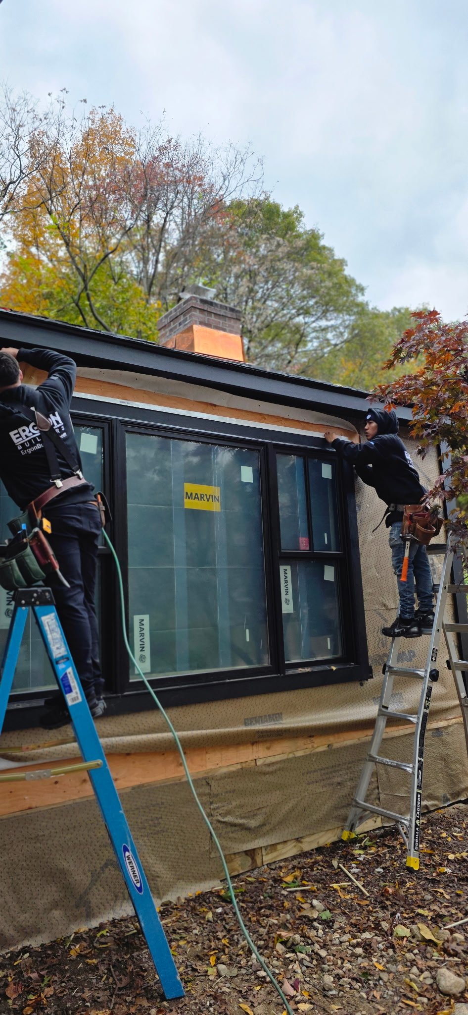 Two construction workers installing a large window on a house. One worker is on a blue ladder, the other on a step ladder, working on the window frame. The house is in an outdoor setting with trees showing autumn foliage.