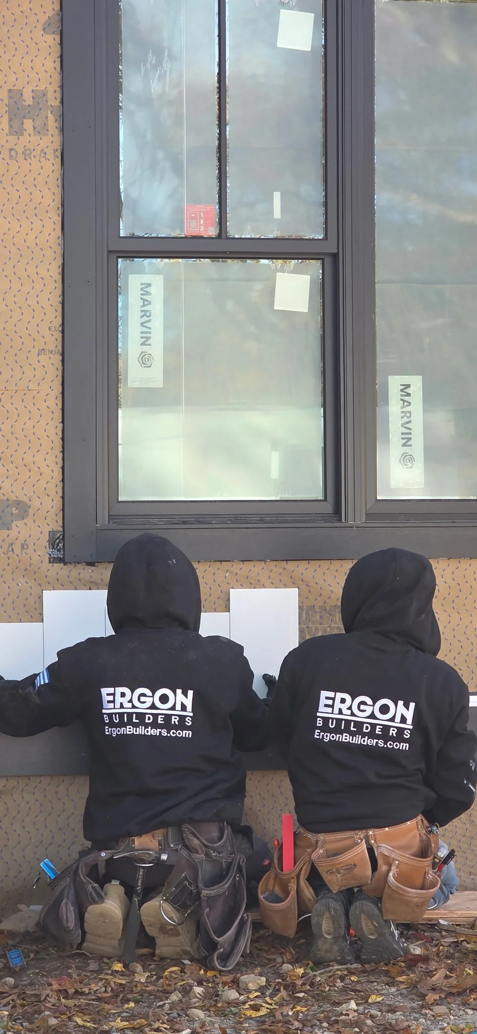 Two construction workers wearing black hooded jackets with 'ERCON BUILDERS' printed on the back are installing a window. They are kneeling on the ground, which is covered in leaves and debris, with tool belts around their waists. The window is partia