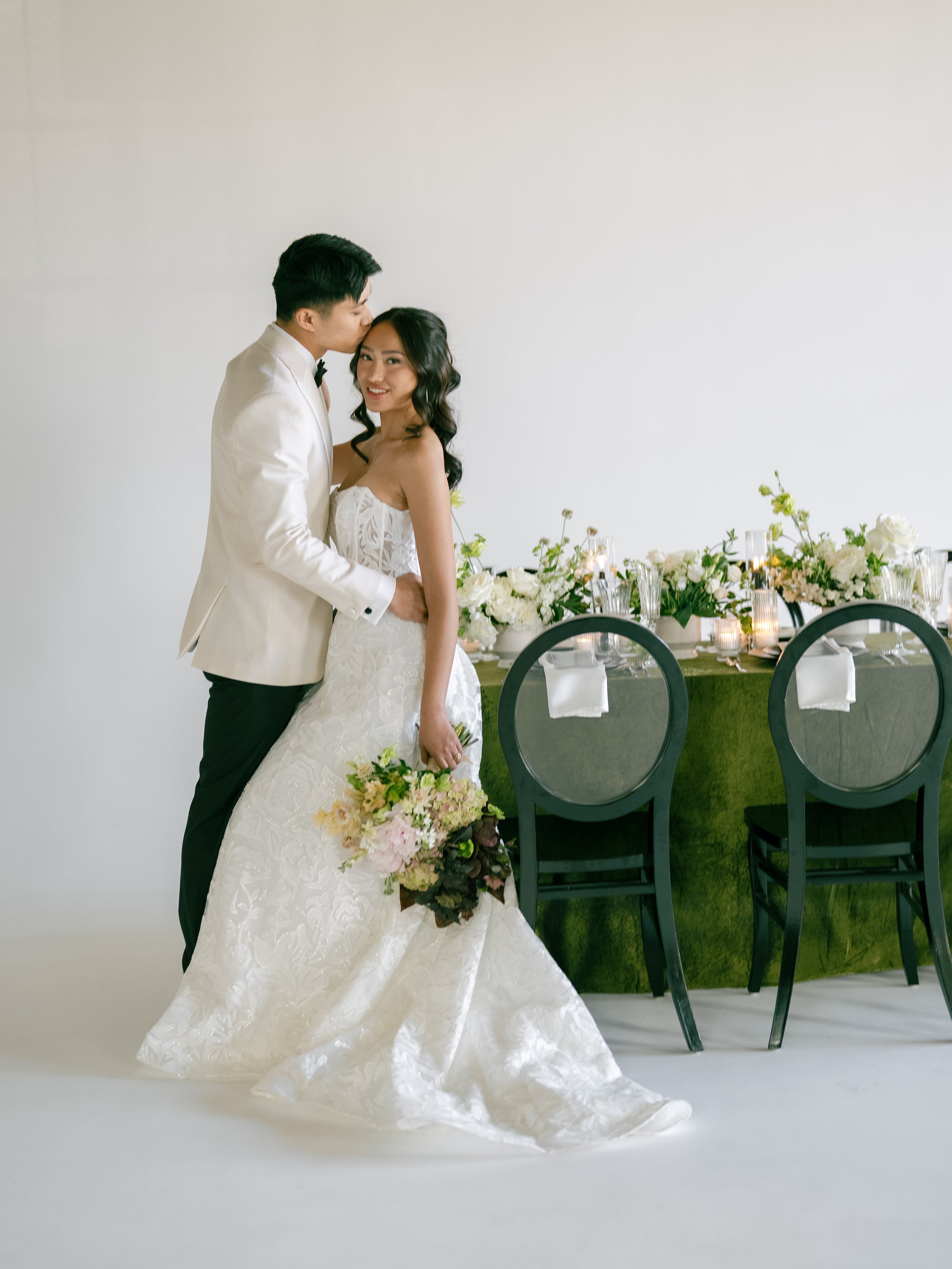 A newlywed couple, a man in a white tuxedo and a woman in a white wedding gown, stands close together in a photo studio with a white background and a decorated table with flowers and candles behind them.