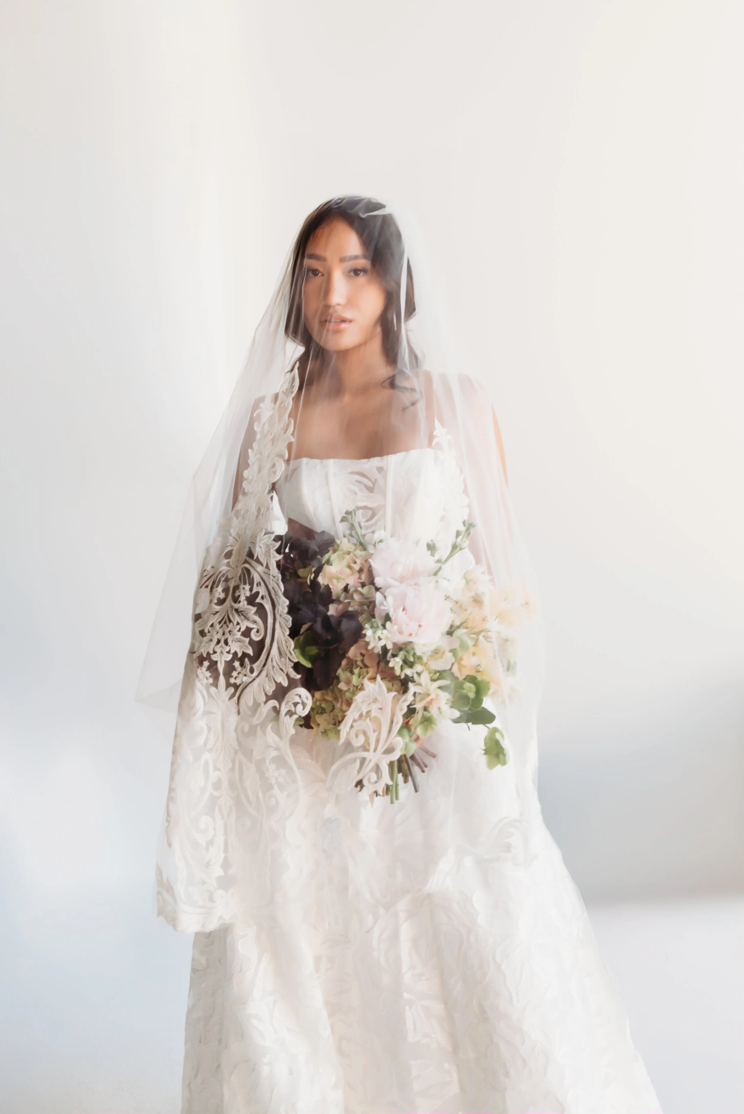 A woman in a white wedding dress with lace details, wearing a veil, and holding a large bouquet of light pink, dark purple, and white flowers, standing against a plain white background.