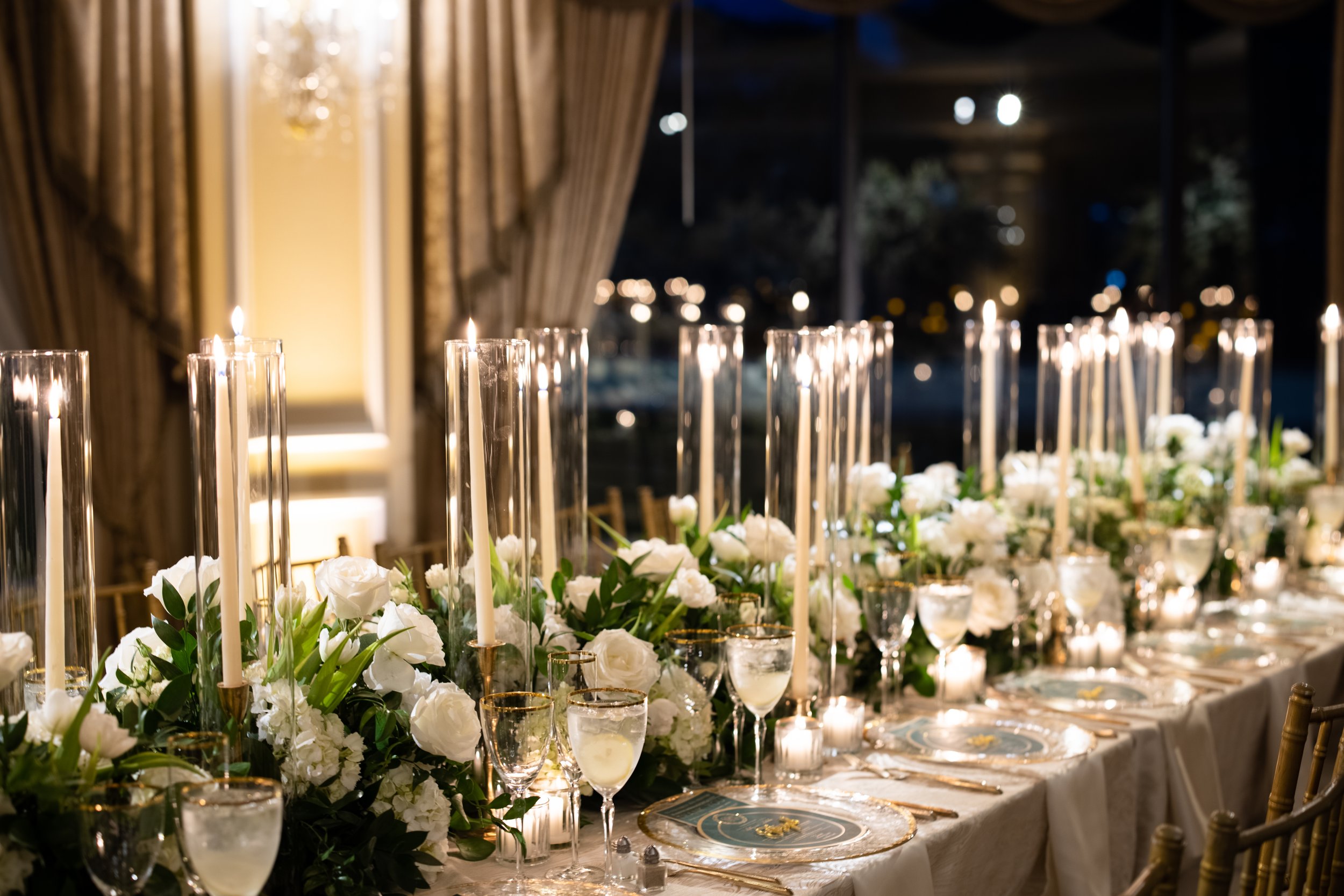 Elegant banquet table decorated with white flowers, tall candles in glass holders, and fine china, set in a warmly lit room with draped curtains and a nighttime view through windows.