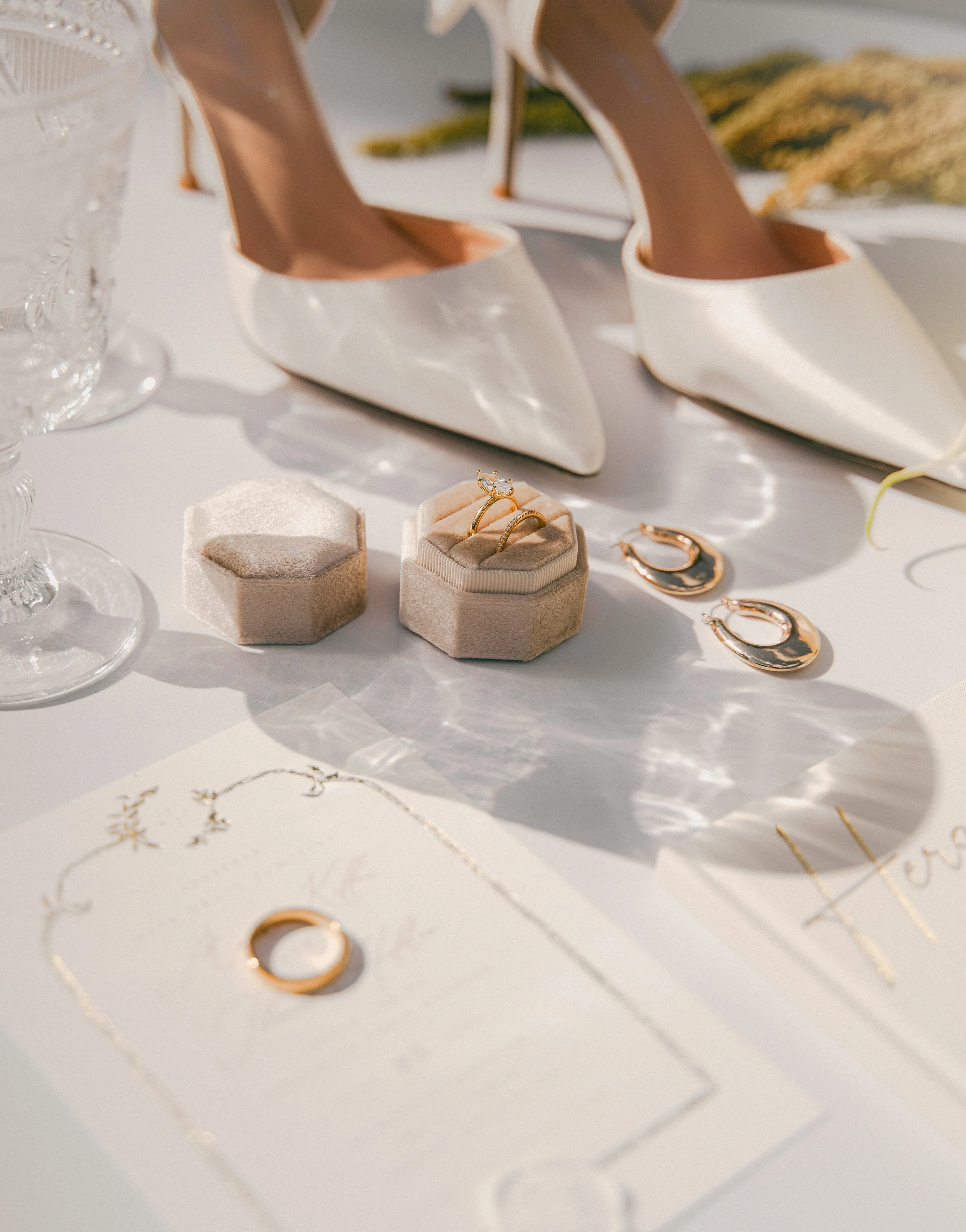 Wedding jewelry including rings in a velvet box, gold hoop earrings, and a gold ring on a table with white high-heeled shoes, a glass, and a wedding invitation.