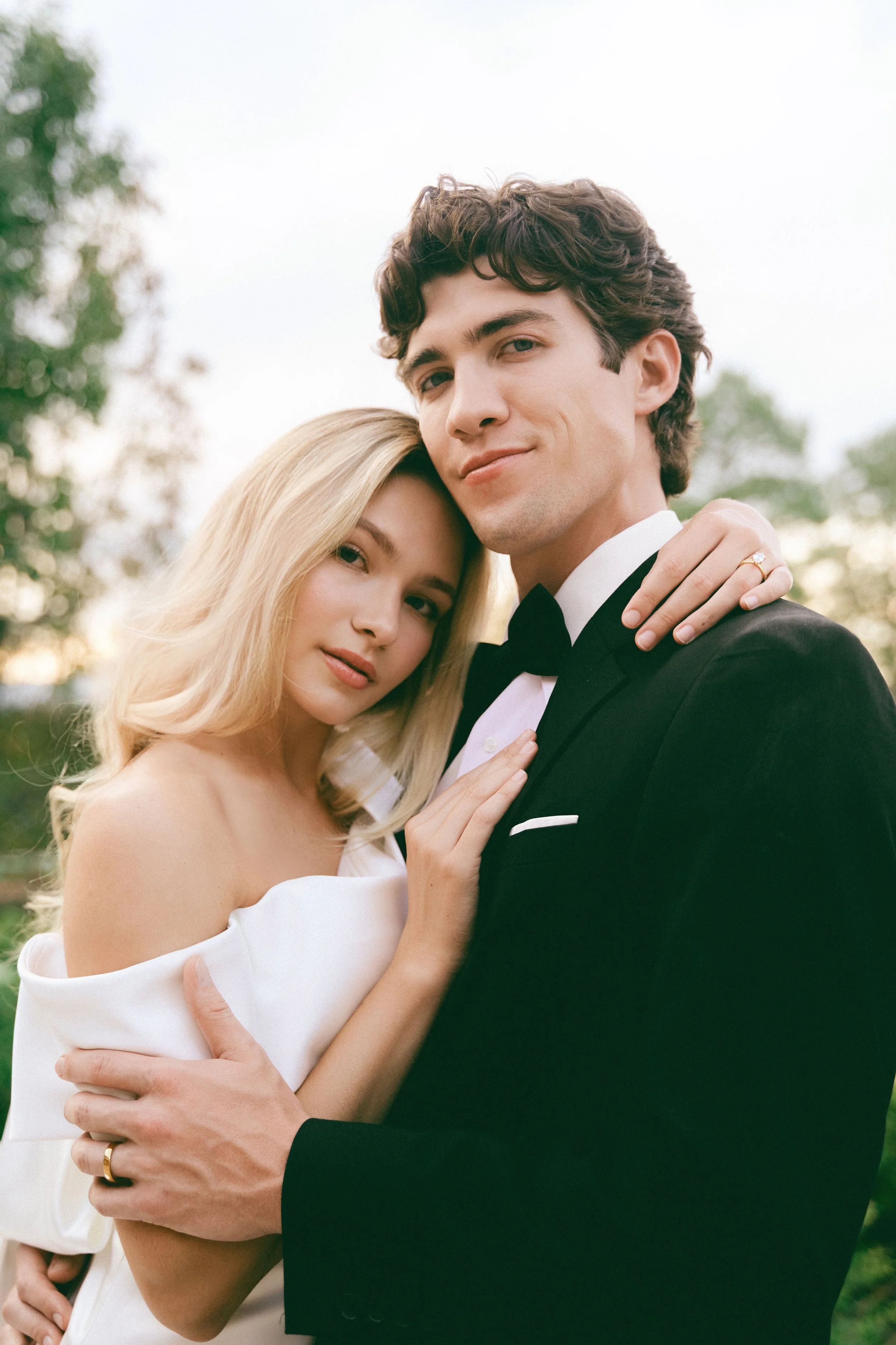 A young couple dressed in wedding attire, with the woman in a white off-shoulder dress and the man in a black tuxedo, posing outdoors during sunset.