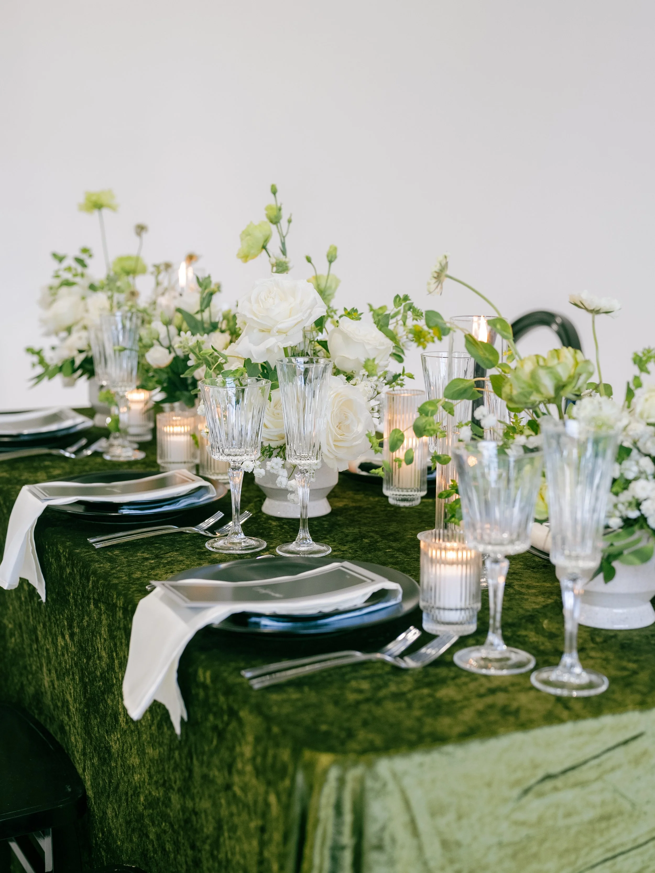 Elegant table setting with white flowers, candles, and glassware on a dark green tablecloth for a special event.