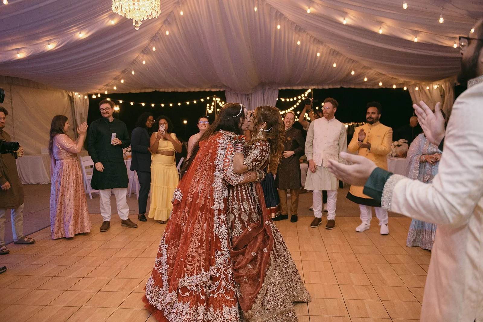 Two women dressed in colorful traditional Indian wedding attire sharing a kiss on the dance floor at a wedding reception, surrounded by friends and family under a decorated tent.