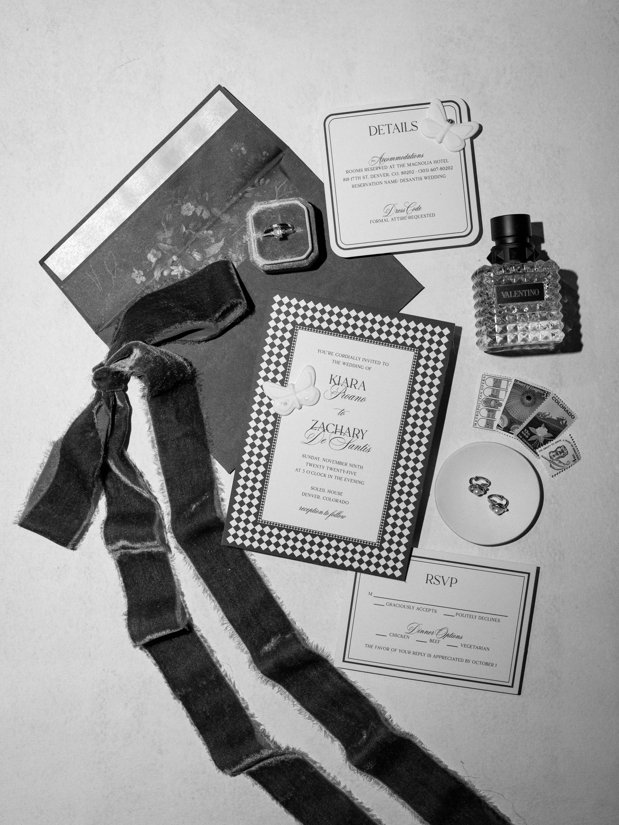 Wedding invitation suite with RSVP card, details card, and invitation for Kiara and Zachary's wedding, along with wedding rings in a box, a perfume bottle, postage stamps, and a ribbon, arranged on a flat surface.