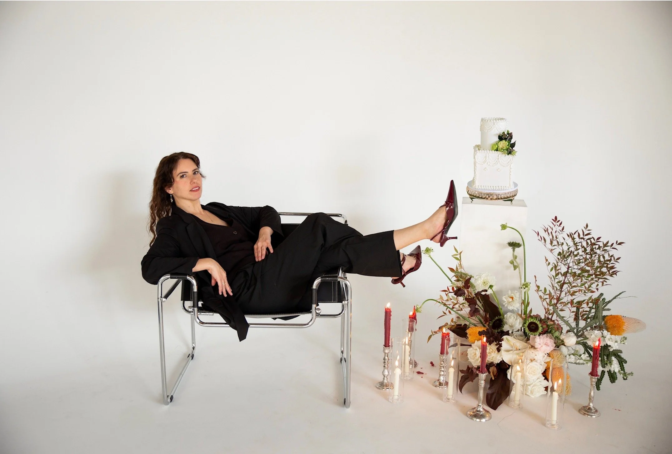 Woman in a black suit lying on a modern chair with her legs raised, wearing high heels, next to a decorated wedding cake surrounded by candles and flowers against a plain white background.