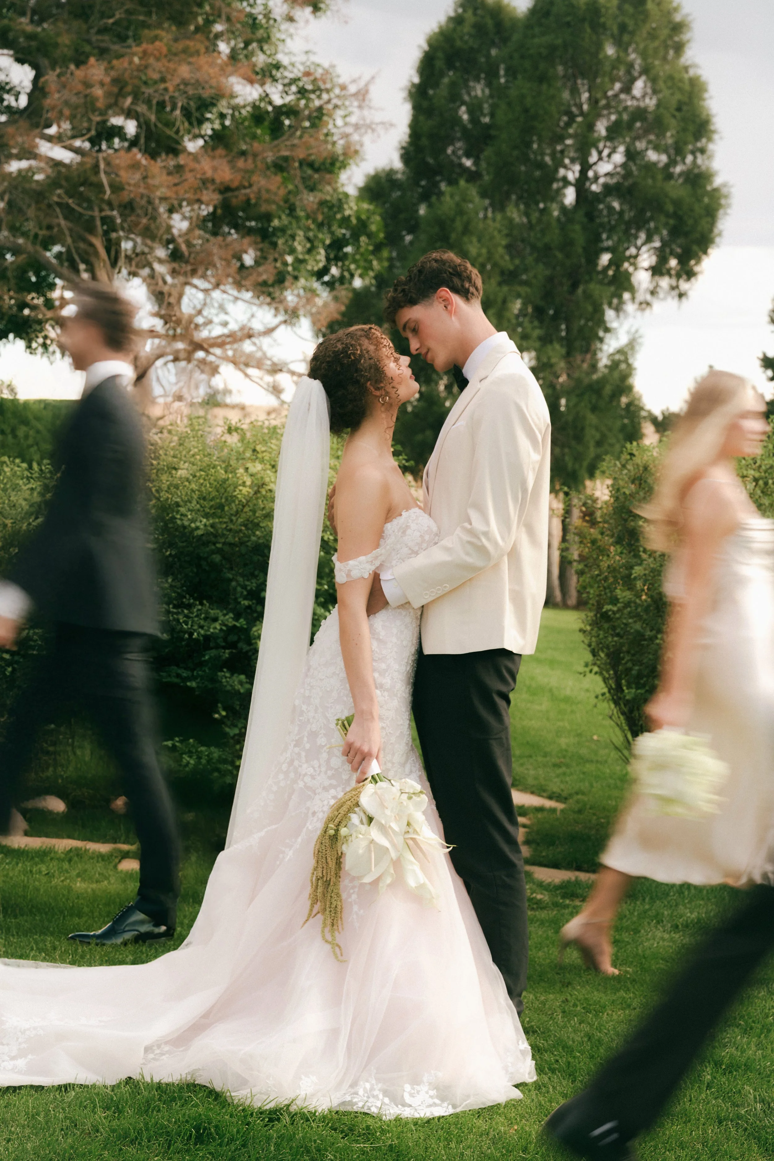 A bride and groom stand close together outdoors, celebrating their wedding, surrounded by blurred guests walking by.