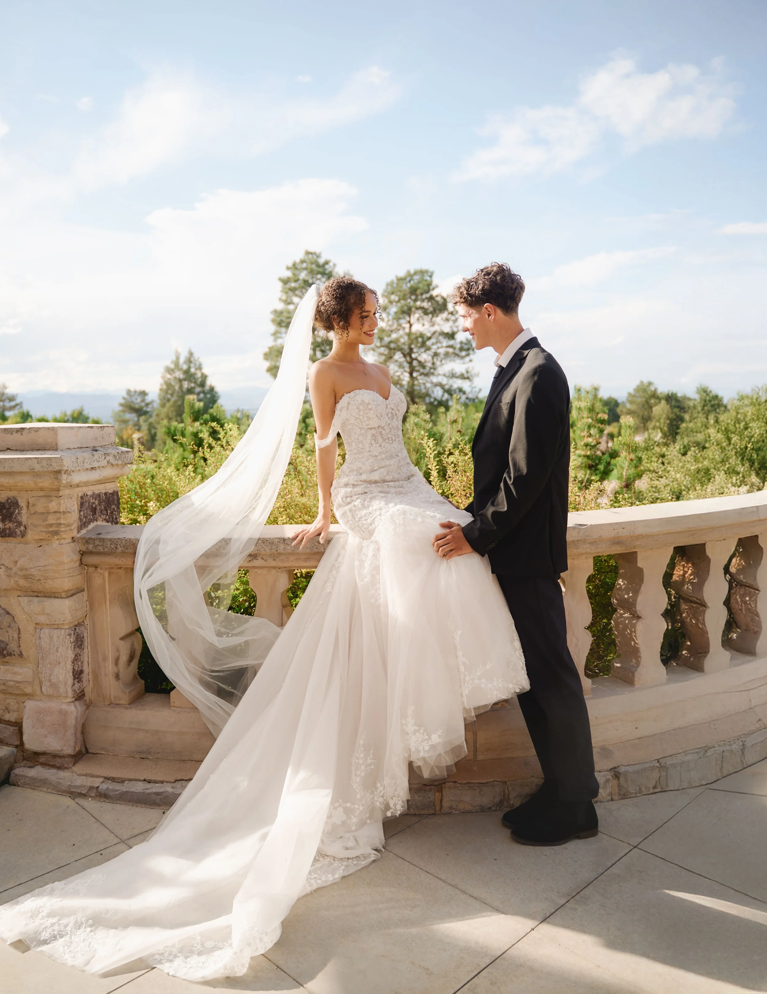 A bride and groom outdoors on their wedding day. The bride is sitting on a stone ledge wearing a white lace wedding gown with a long veil, while the groom stands facing her, wearing a black tuxedo. They are smiling at each other with a backdrop of trees and a partly cloudy sky.