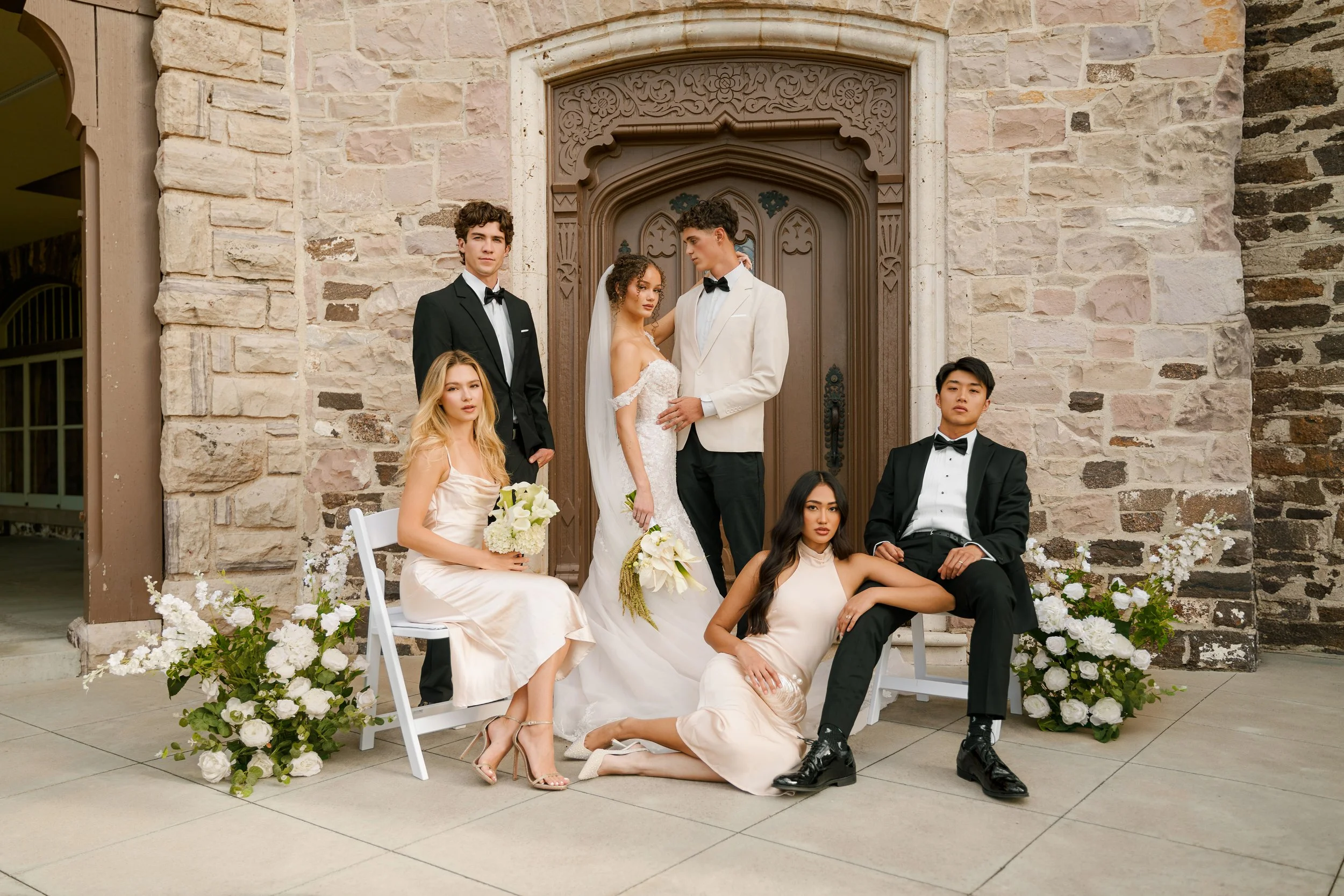 A group of seven people dressed in wedding attire, posing in front of a stone building with a wooden door, surrounded by white floral arrangements.