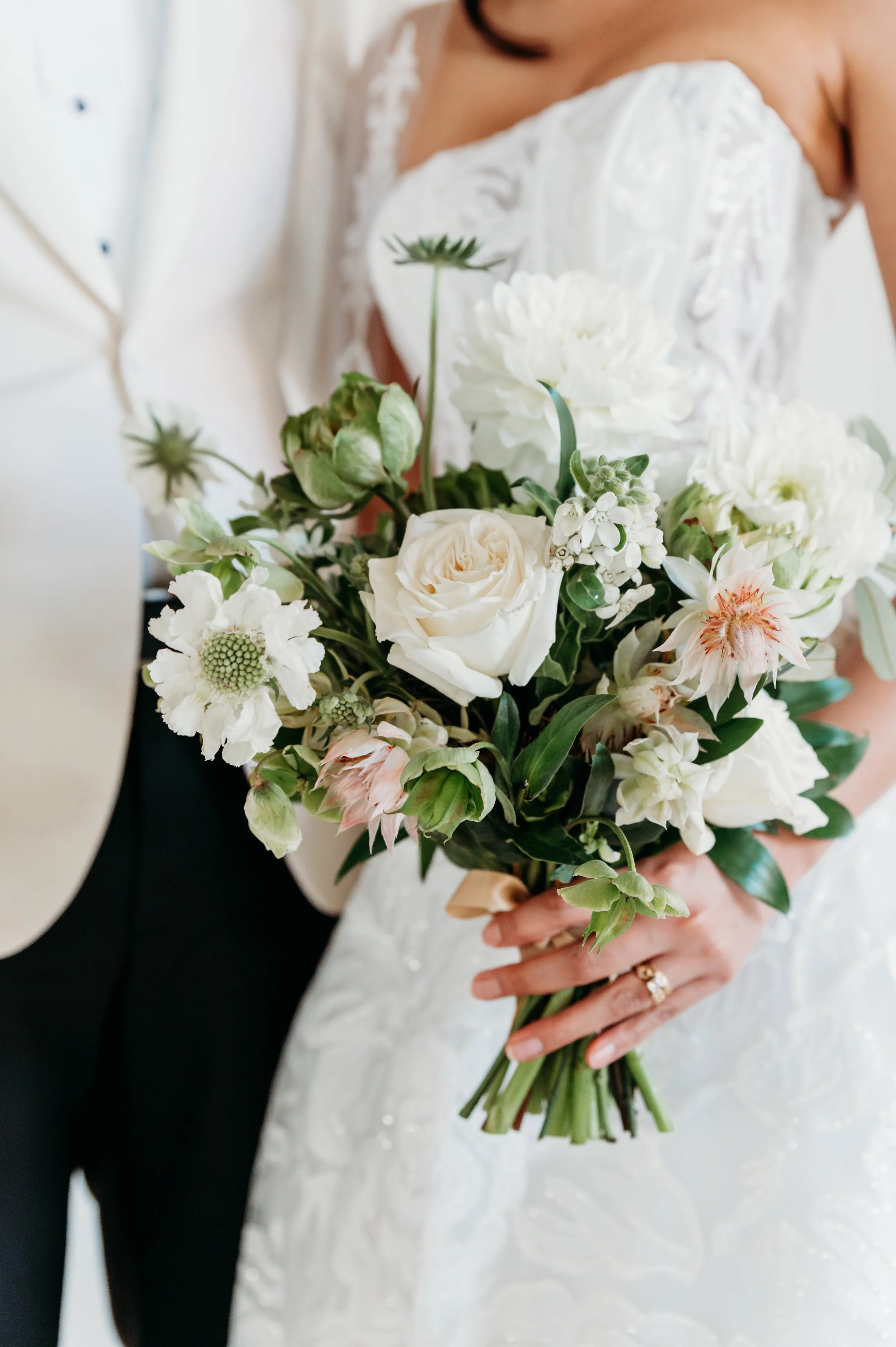 Bride holding a bouquet of white and light pink flowers, wearing a white wedding dress with lace details.