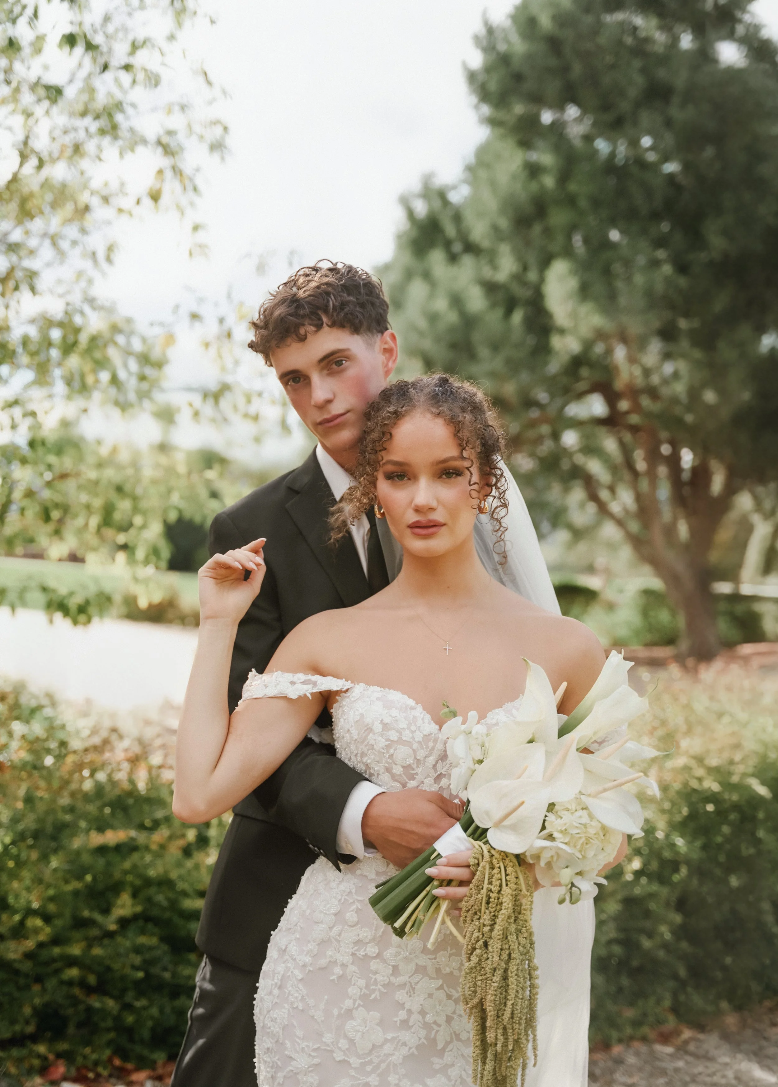 A newlywed couple standing outdoors, with the man in a black suit and the woman in a white wedding dress holding a bouquet of white flowers, under a background of trees and greenery.