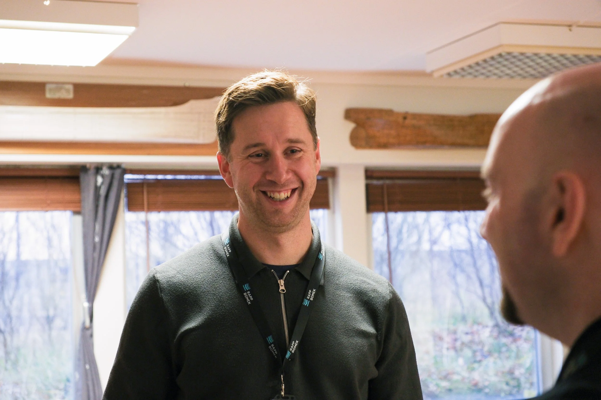 Two men smiling and talking indoors near windows with brown blinds and curtains
