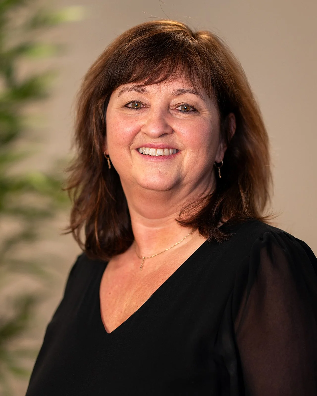 A woman with shoulder-length brown hair smiling, wearing a black top with sheer sleeves, earrings, and a gold necklace, standing indoors with a neutral background.