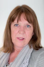 Close-up of a middle-aged woman with shoulder-length brown hair, light skin, blue eyes, and wearing a light gray top, standing against a plain background.