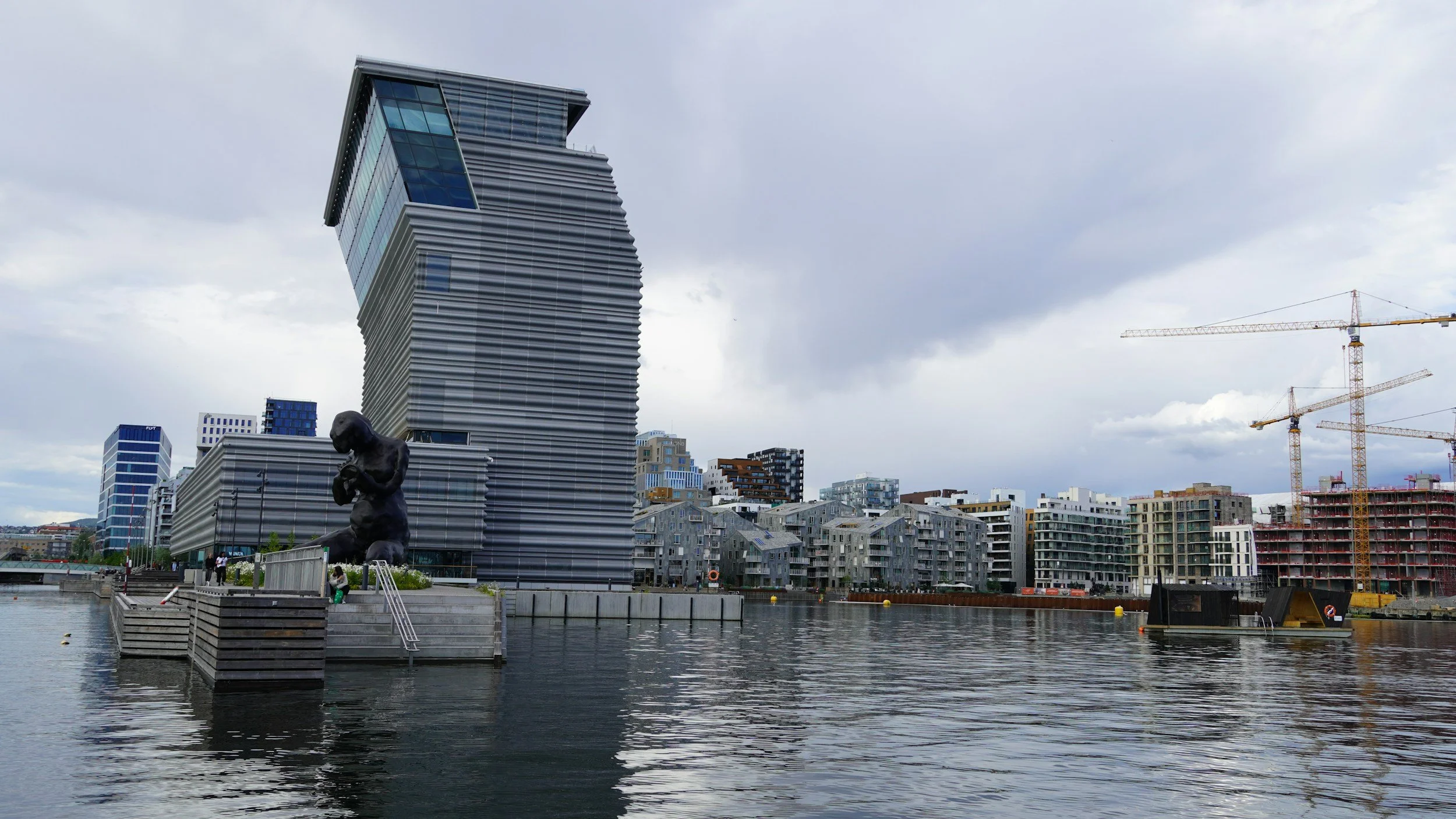 Modern cityscape with a distinctive tilted skyscraper, a large black sculpture of a praying person near water, and construction cranes in the background under a cloudy sky.