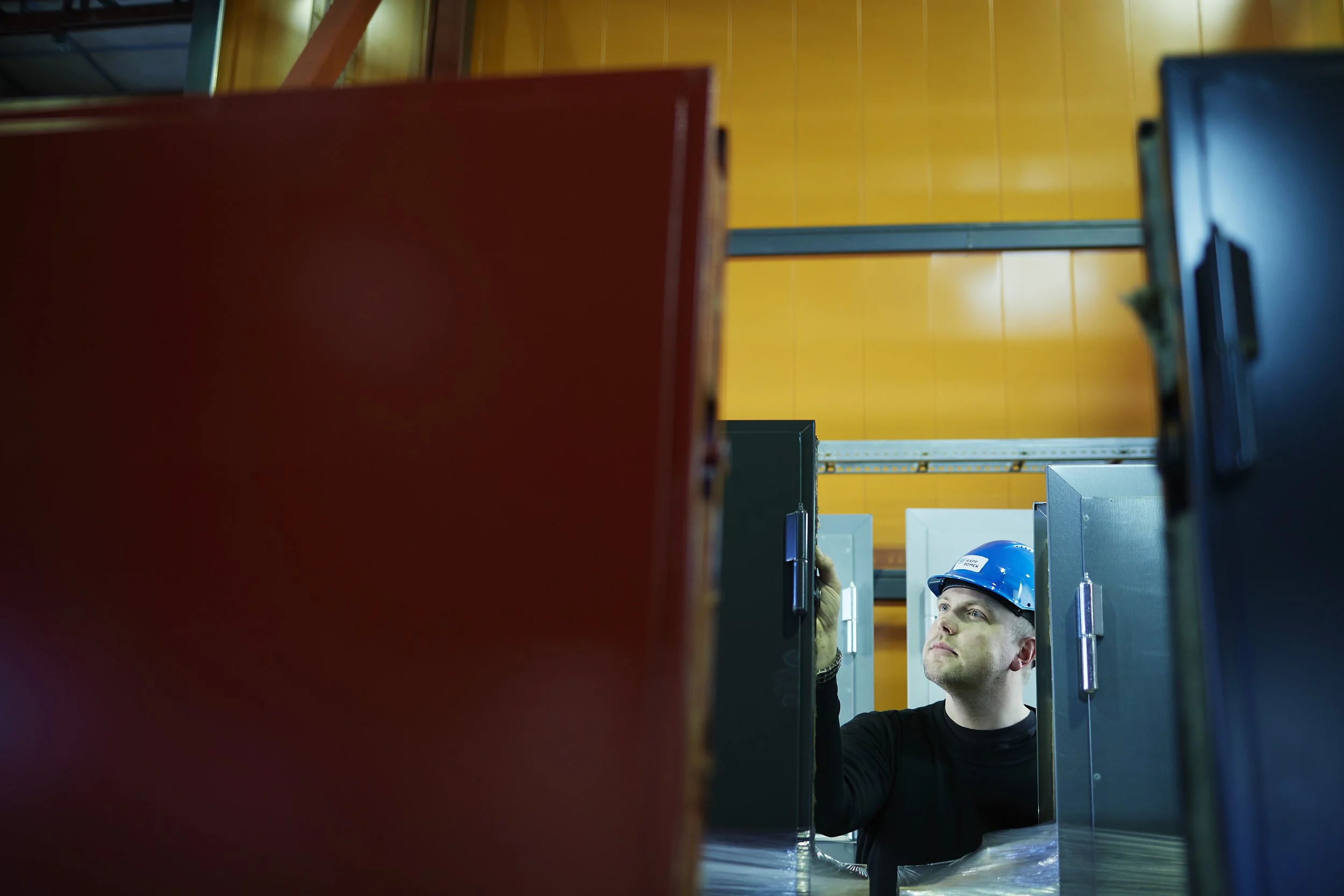 A worker in safety gear, including a blue helmet, inspecting electrical panels inside an industrial facility with yellow and orange walls.