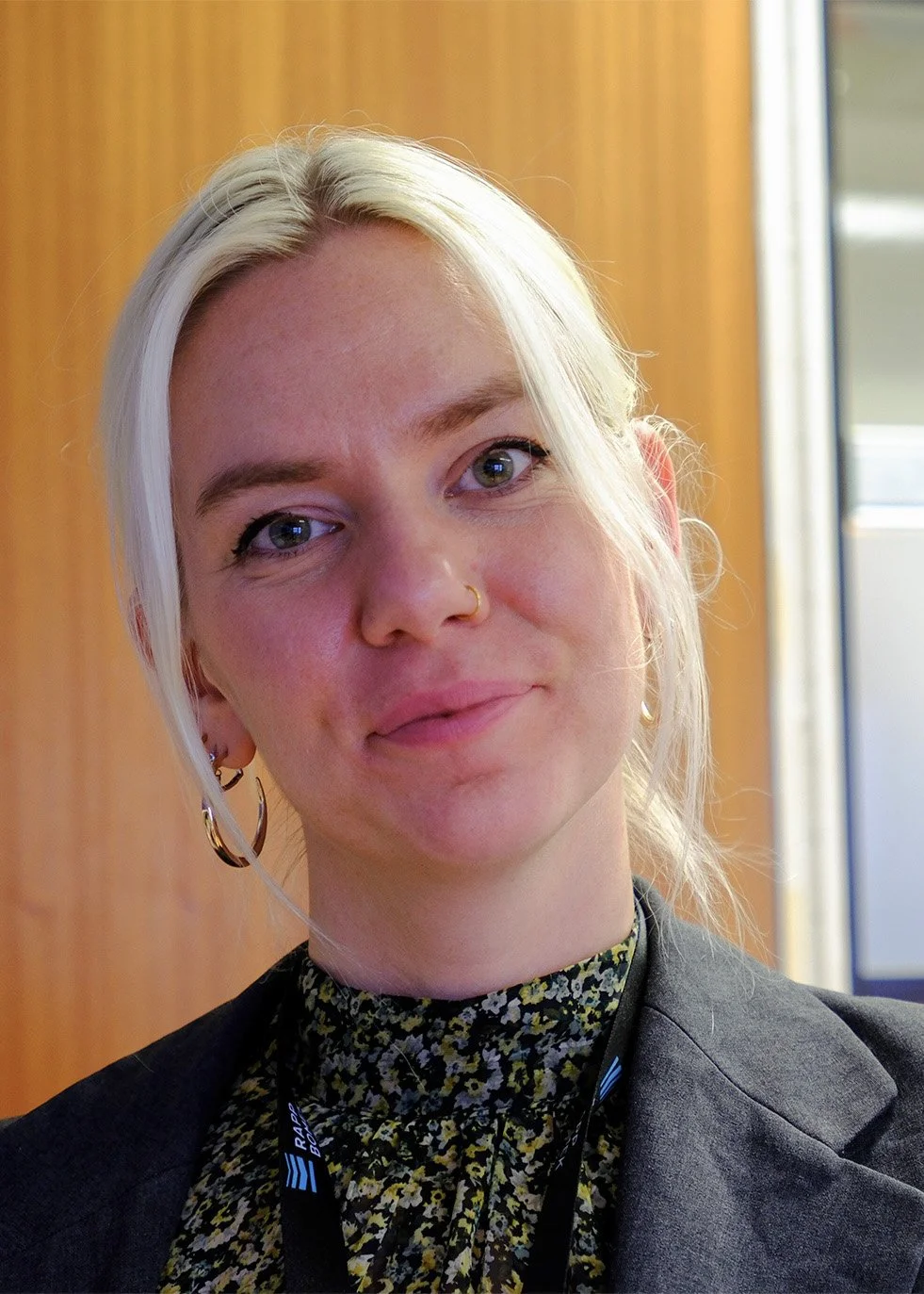 Close-up of a woman with blonde hair, nose ring, earrings, and a floral blouse, smiling softly indoors.