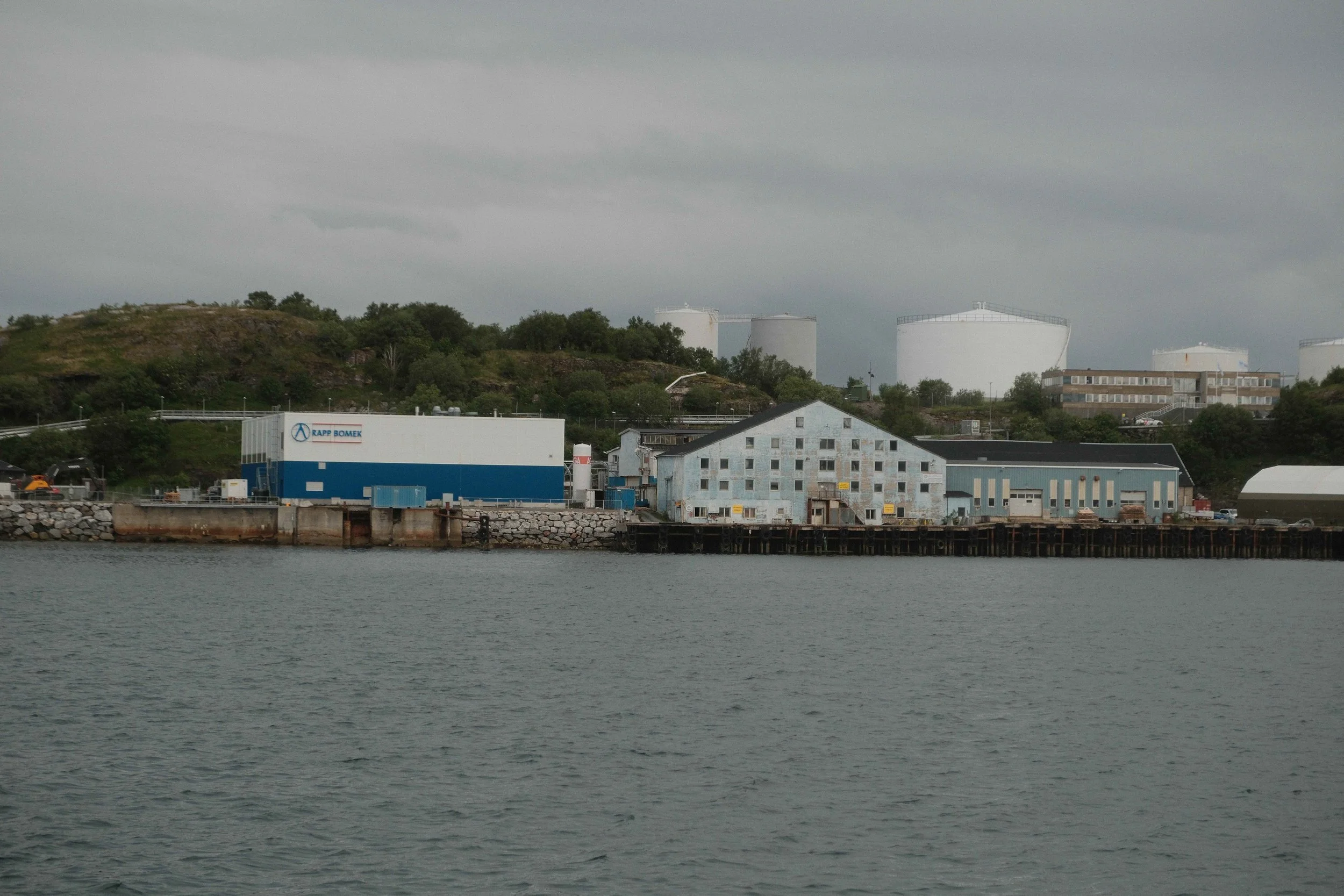 View of industrial buildings along a waterway, with large white storage tanks in the background and a cloudy sky.