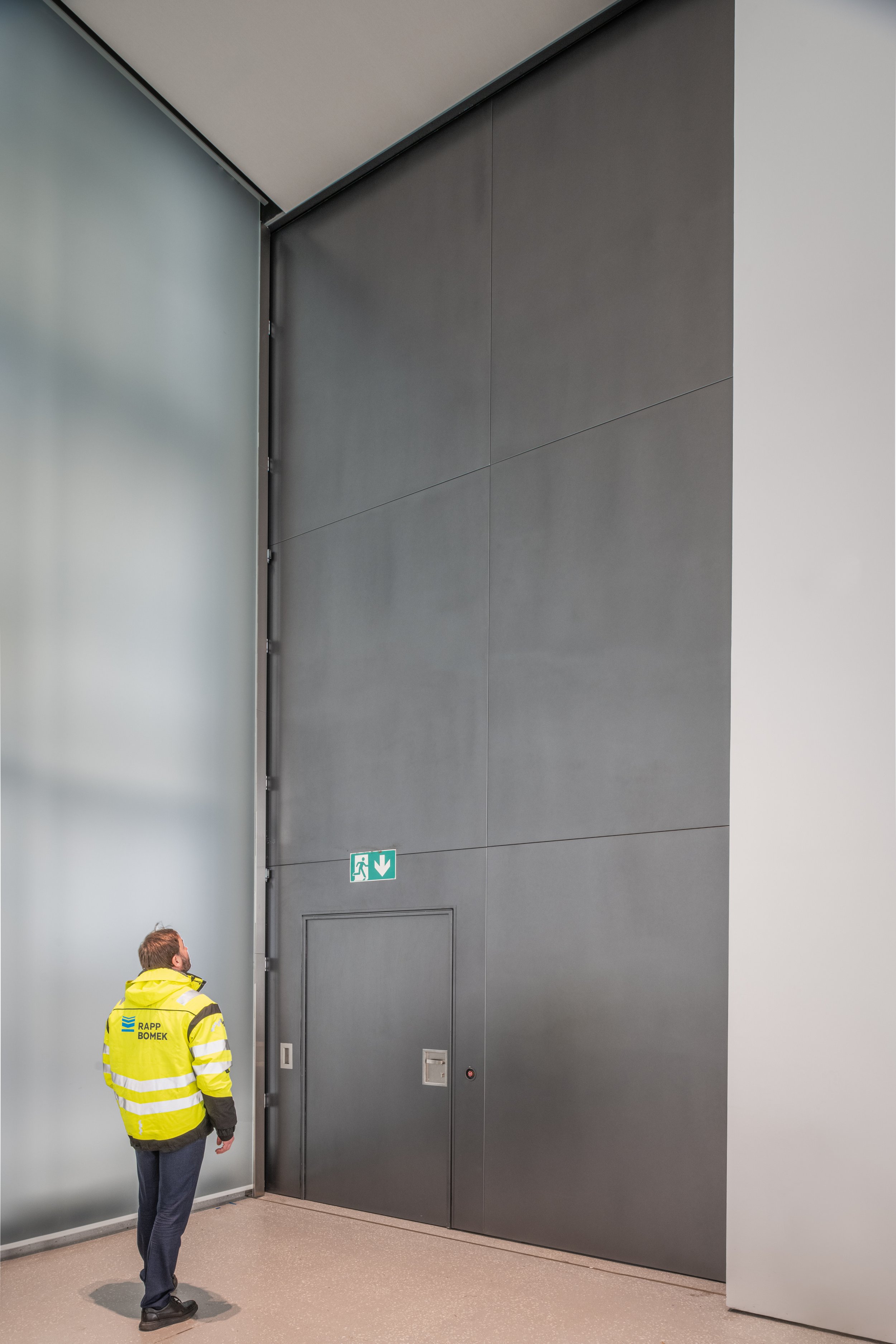 A man in a yellow safety jacket stands inside a large modern building near a gray wall with an emergency exit sign and a small door.