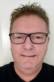 A middle-aged man with short, light brown hair and glasses smiling indoors with a plain white background.