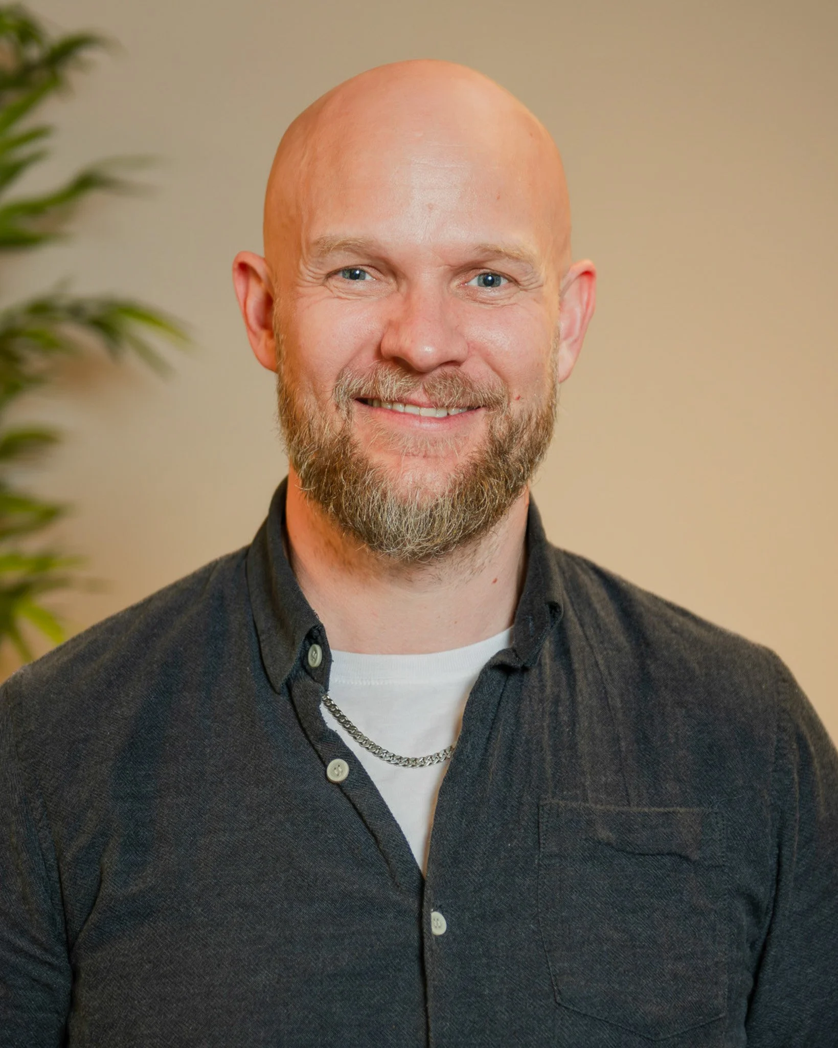 Close-up of a man with a bald head, beard, and mustache, smiling, wearing a dark button-up shirt over a white t-shirt, with a chain necklace, standing in front of a plain background.