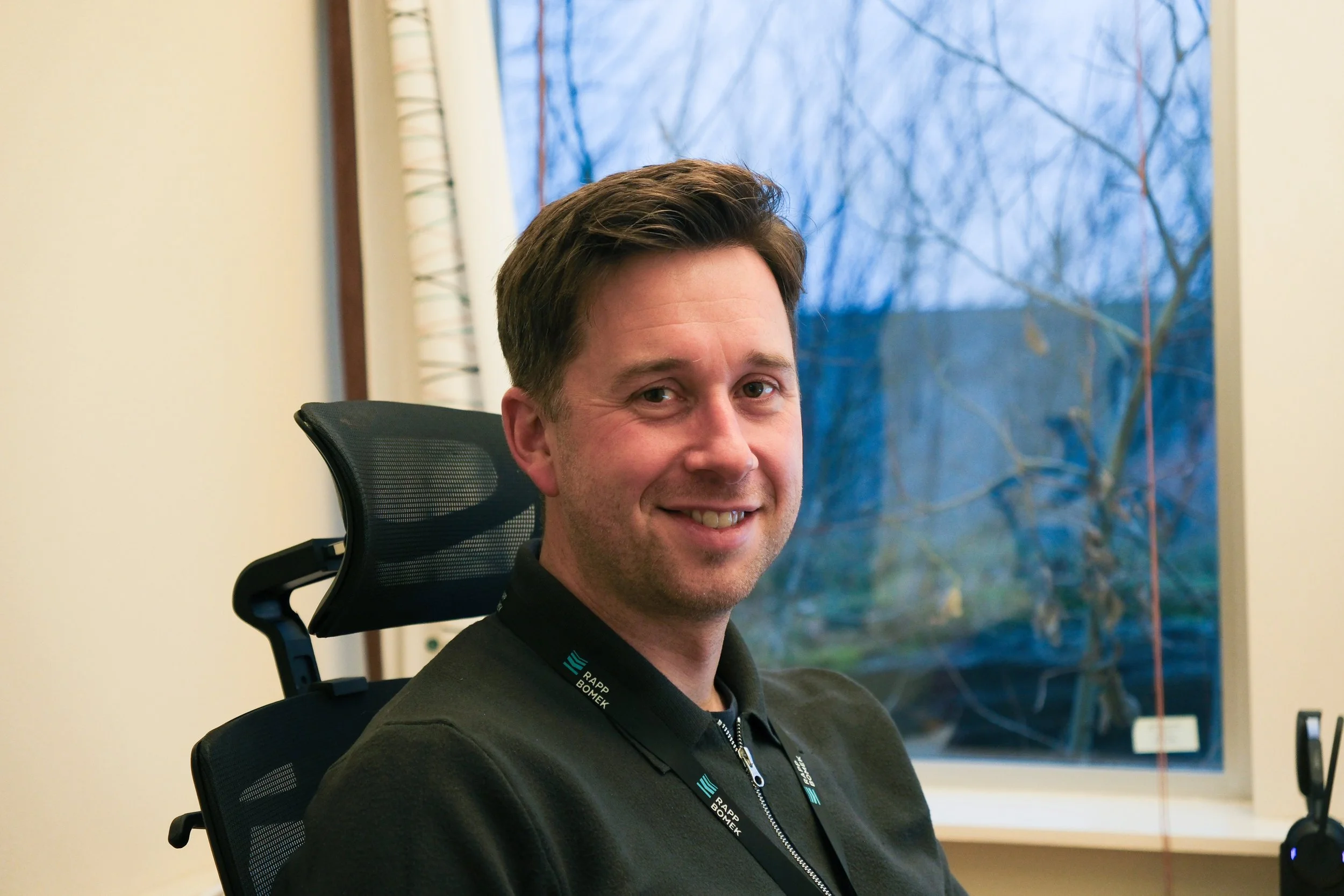 A smiling man sitting in an office chair in front of a window with trees outside.