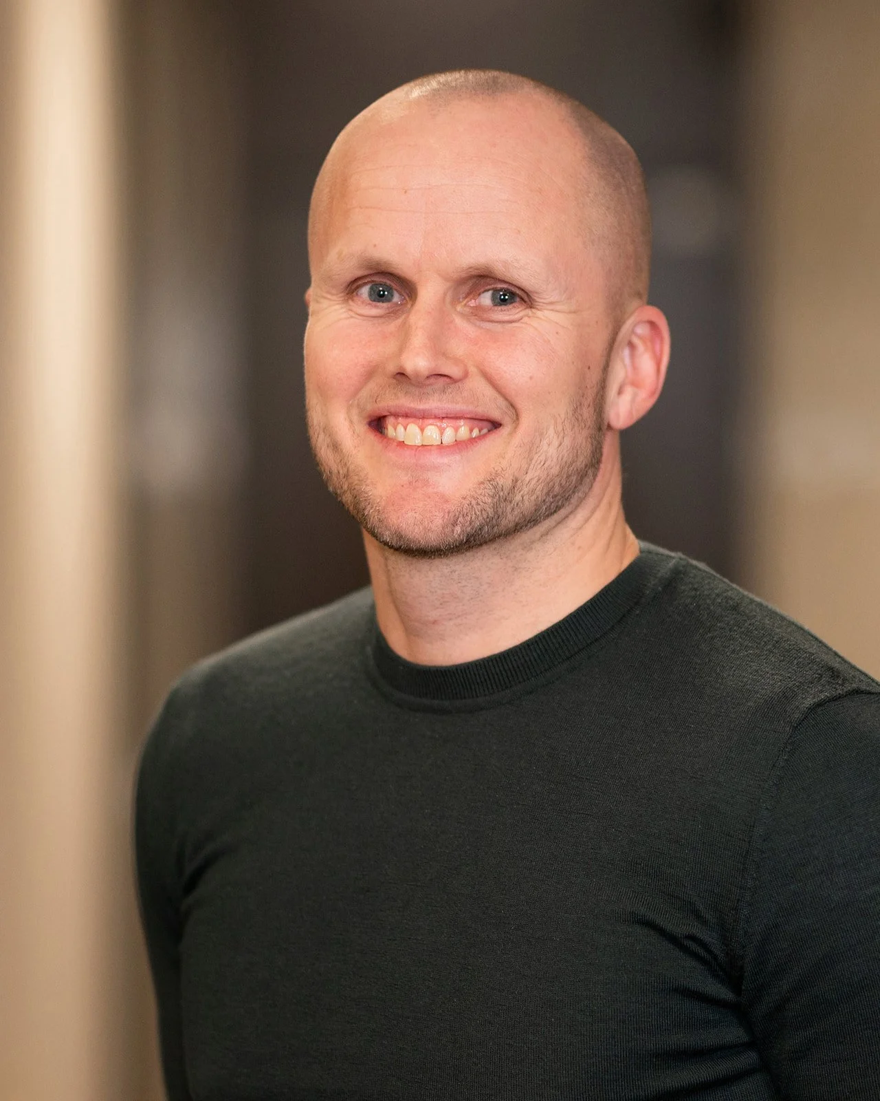 A smiling man with a shaved head and stubble, wearing a black shirt, standing indoors with a blurred background.