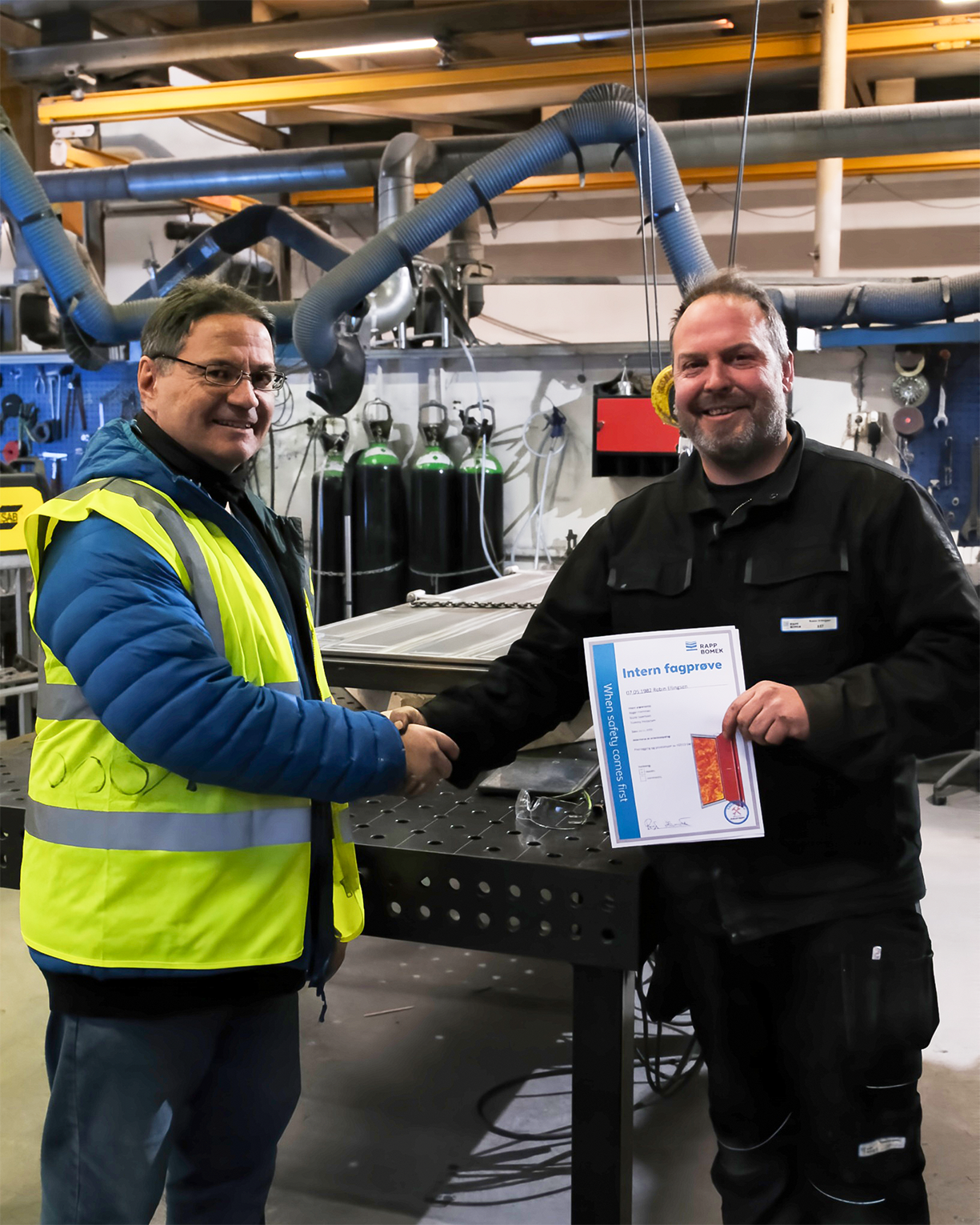 Two men shaking hands in a workshop, one in a high-visibility vest, the other holding a certificate, surrounded by industrial equipment and ventilation pipes.