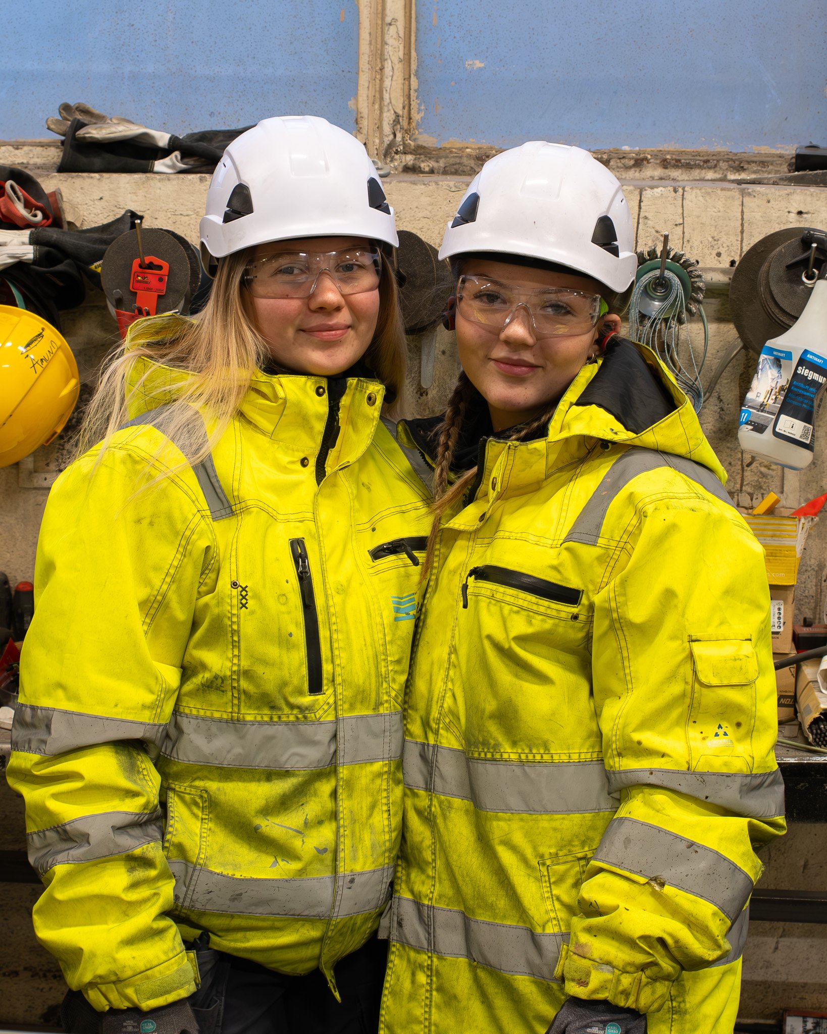 Two women wearing yellow safety jackets, white helmets, and safety glasses standing in a workshop.