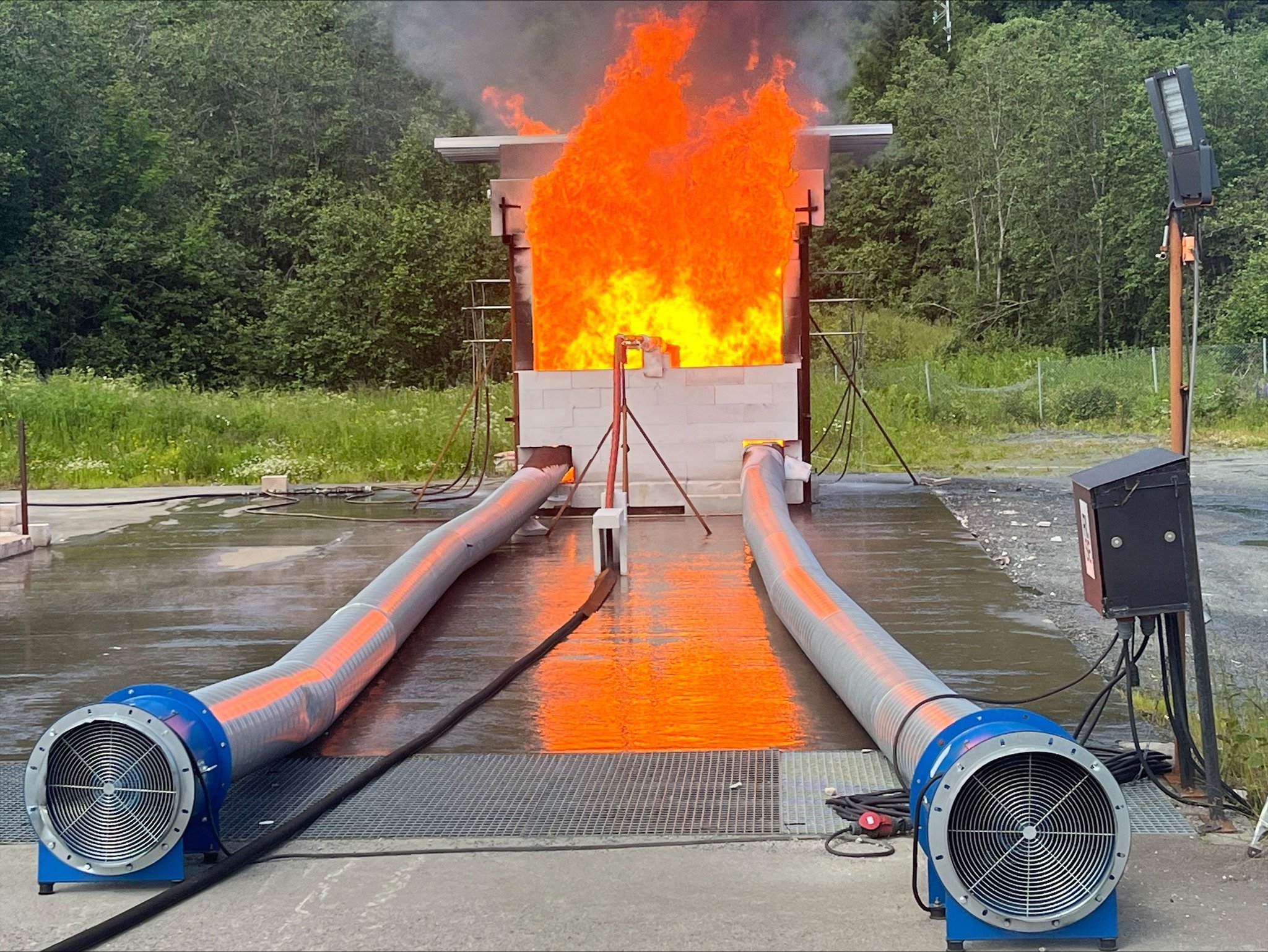 Fire testing setup with flames erupting from a structure, two large flexible pipes connected to fans on the ground, outdoors with trees in the background.