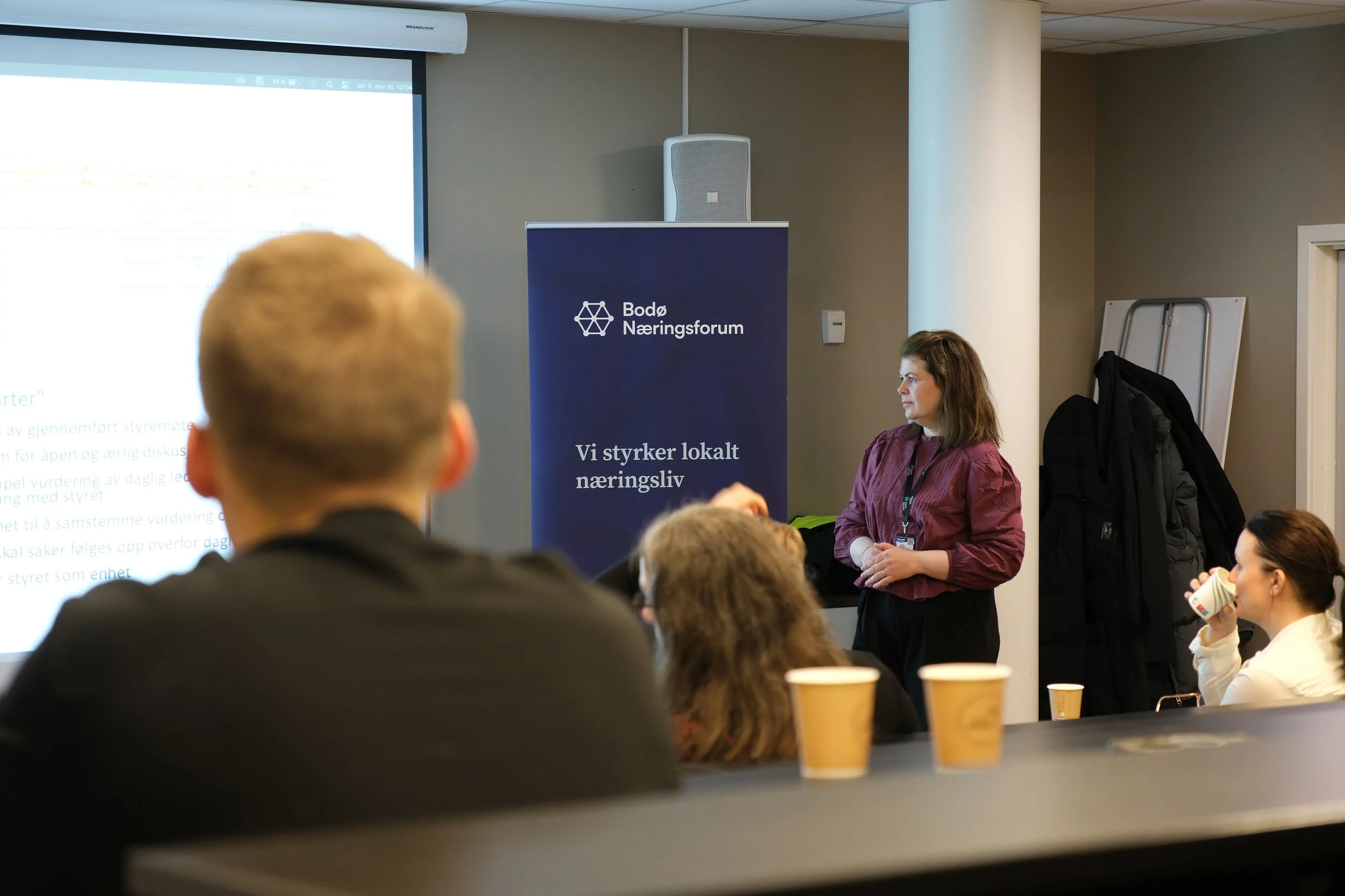 A woman in a maroon shirt presenting at a conference in front of a blue banner with the logo and text of Bodø Næringsforum. Several people are seated and watching her, with some drinking coffee.
