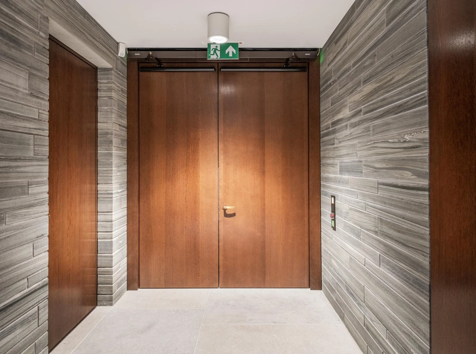 Wooden elevator doors with a green emergency exit sign above and textured gray wall panels on either side.
