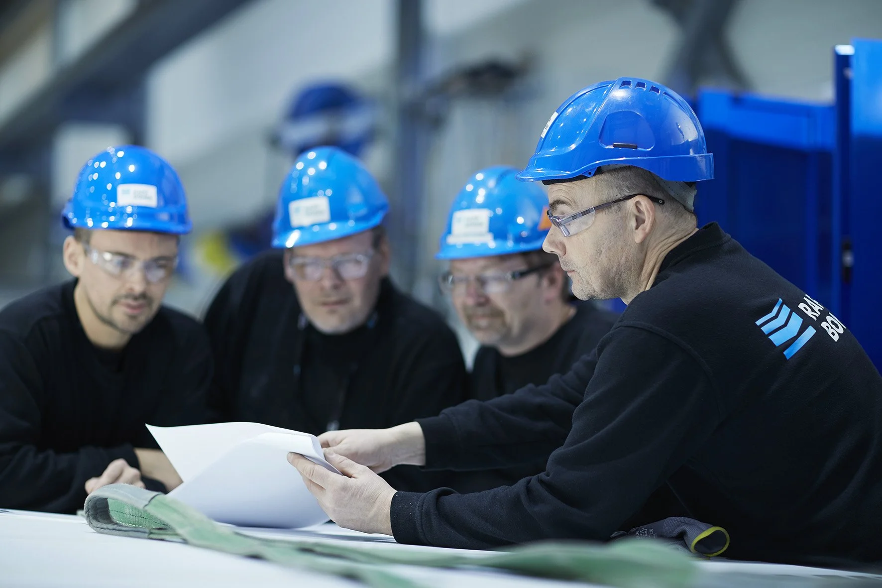 Group of five men wearing blue safety helmets and safety glasses sitting at a table with blueprints, in an industrial or construction setting.