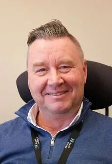 Smiling man with short, styled hair wearing a blue shirt and black lanyard, seated in an office chair against a plain beige wall.
