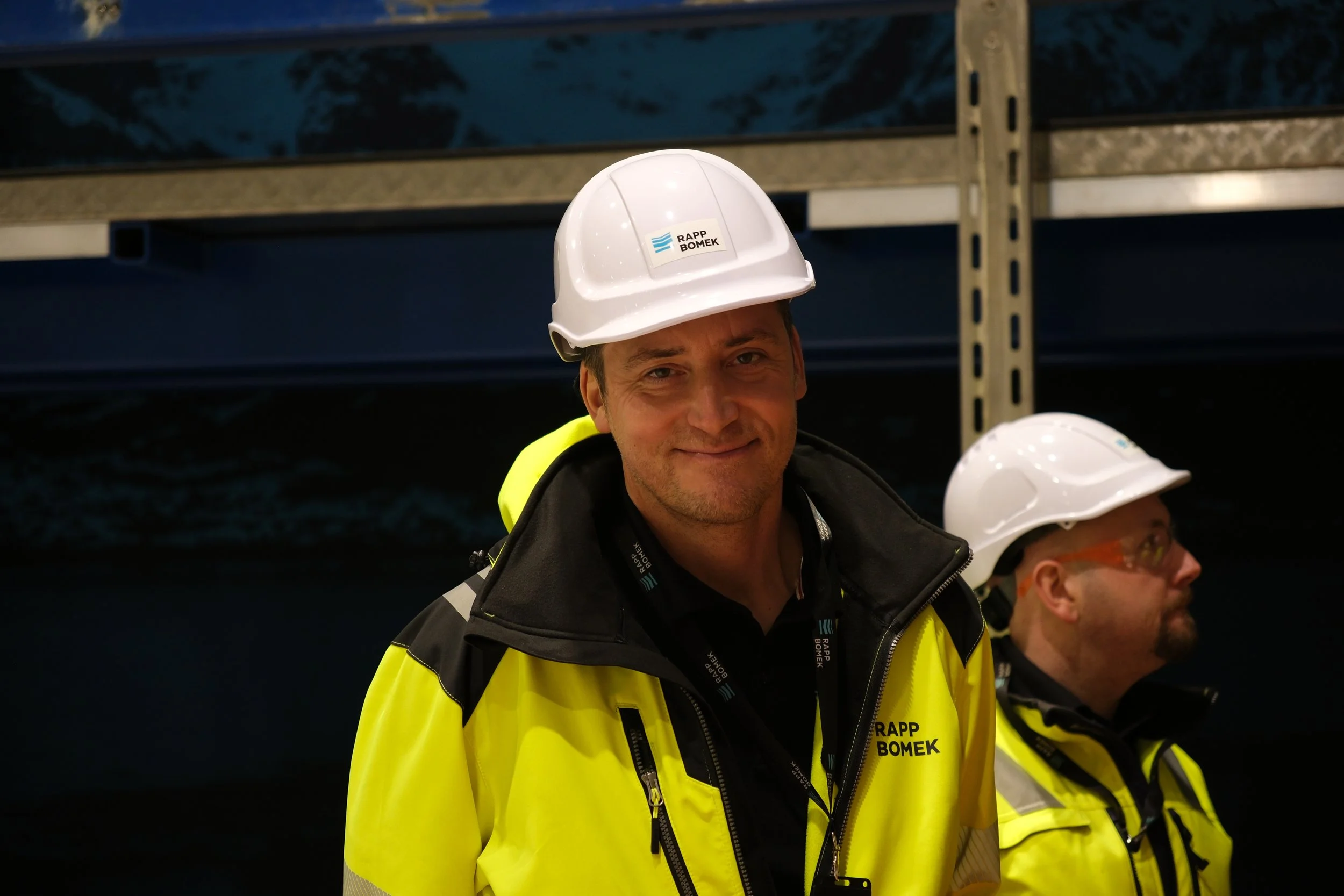 Two men wearing white safety helmets and yellow jacket with black and yellow accents, standing indoors with a dark background and a metal support structure behind them.