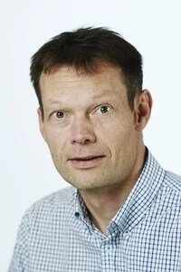 A middle-aged man with short dark hair, wearing a light blue checkered shirt, looking directly at the camera against a plain white background.