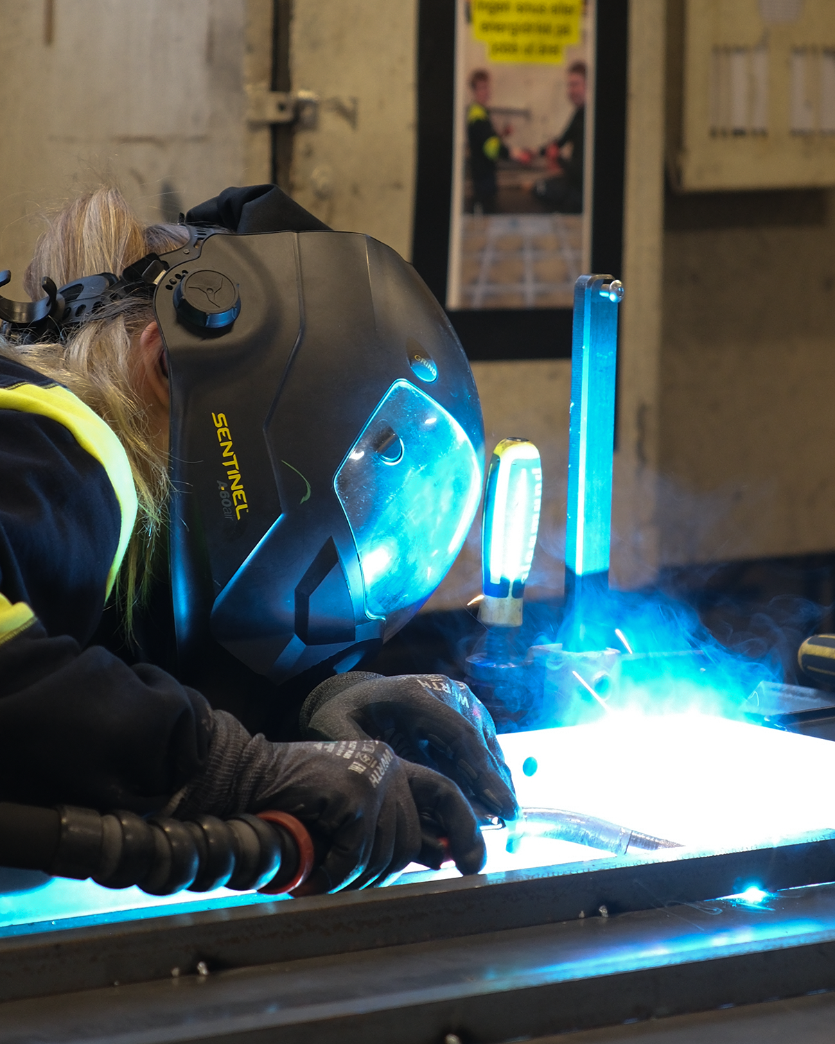A person welding metal with a protective helmet, gloves, and welding equipment; blue sparks and light emitted during welding; workshop setting with a poster on the wall.