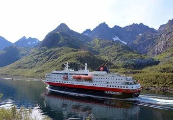 A white and orange cruise ship sailing through calm water with green mountains and rugged peaks in the background.