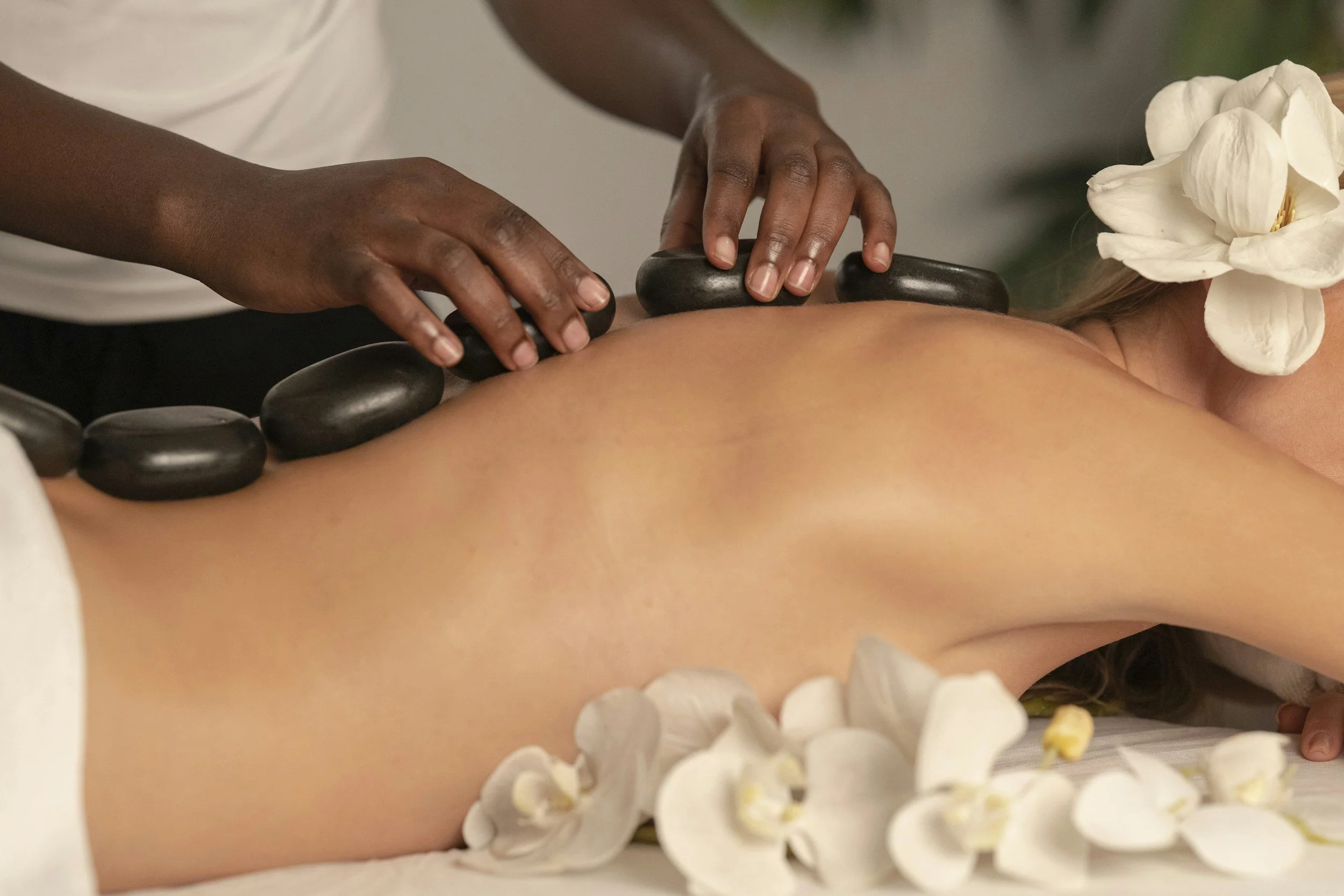 A person receiving a hot stone massage on their back, with their face turned to the side and a white flower near their face. The person is lying on a massage table with white flowers surrounding their body.