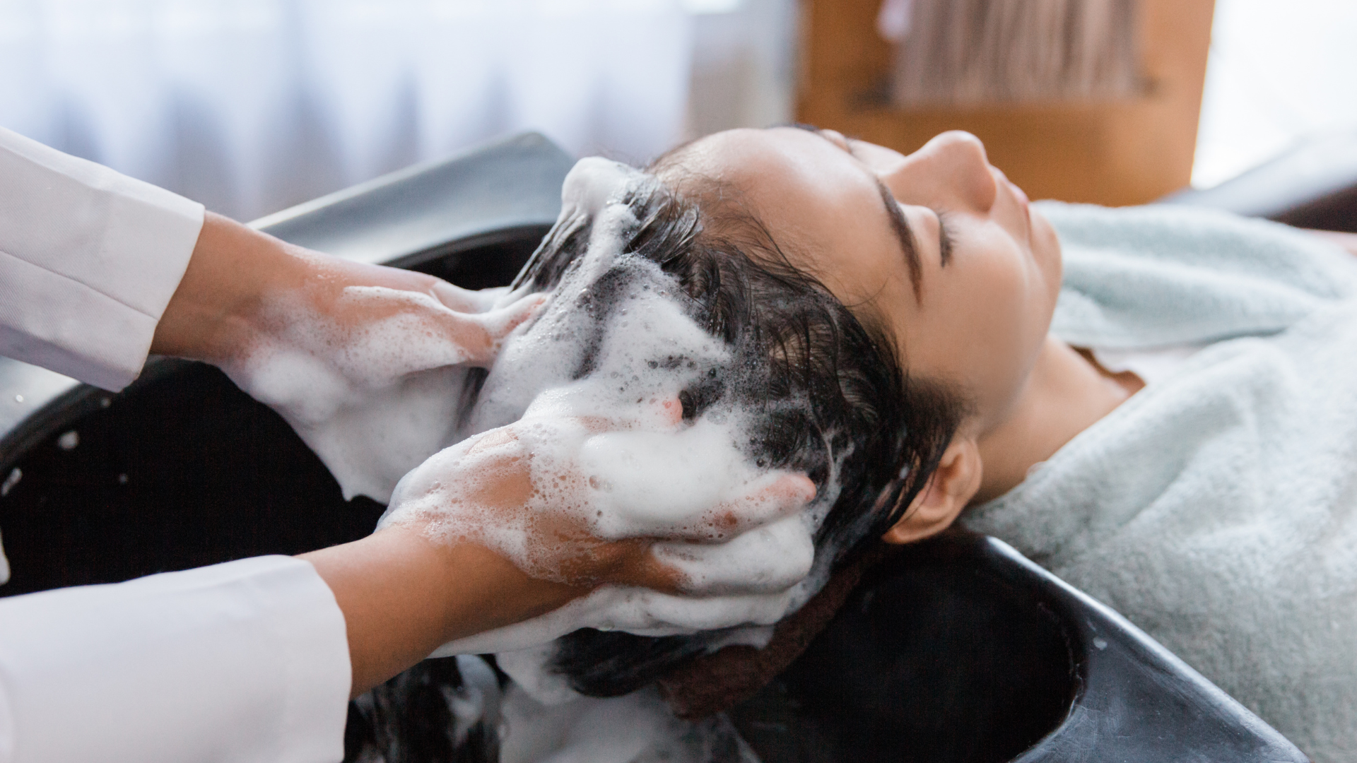 A woman is getting her hair washed at a salon. She is lying back with eyes closed while a stylist washes her hair with soap and water in a black sink.