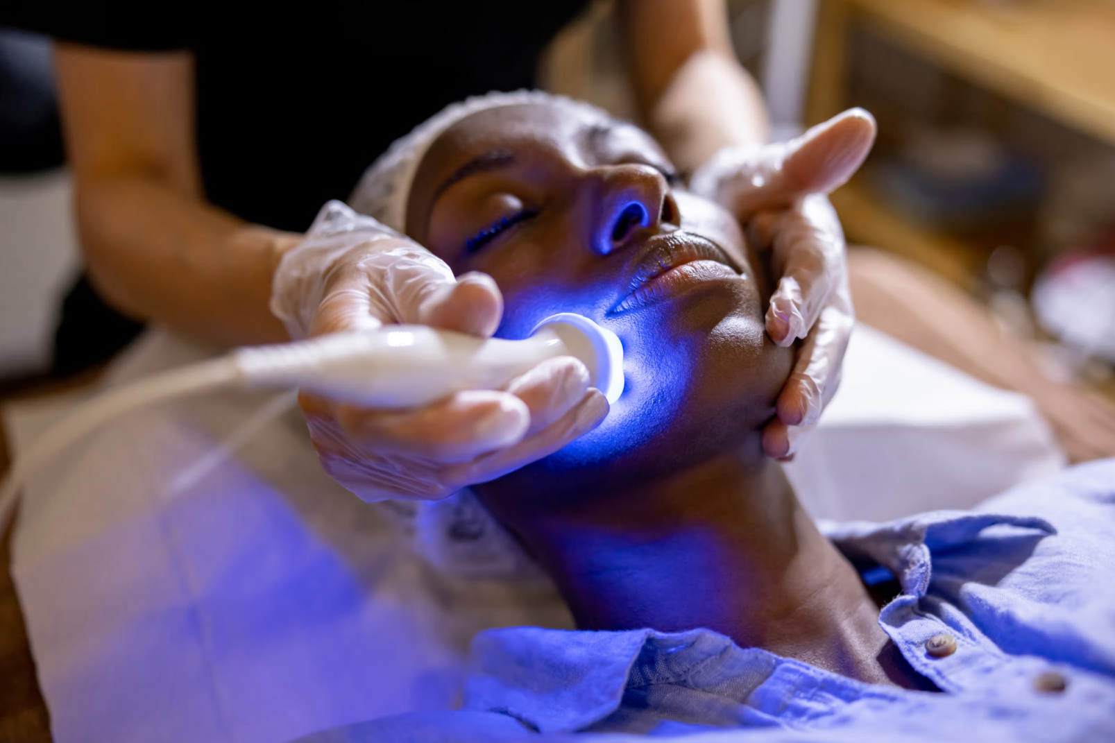 A woman receives a facial treatment with a blue light device in a spa or skincare clinic, lying on a bed, with her eyes closed and a facial mask applied.