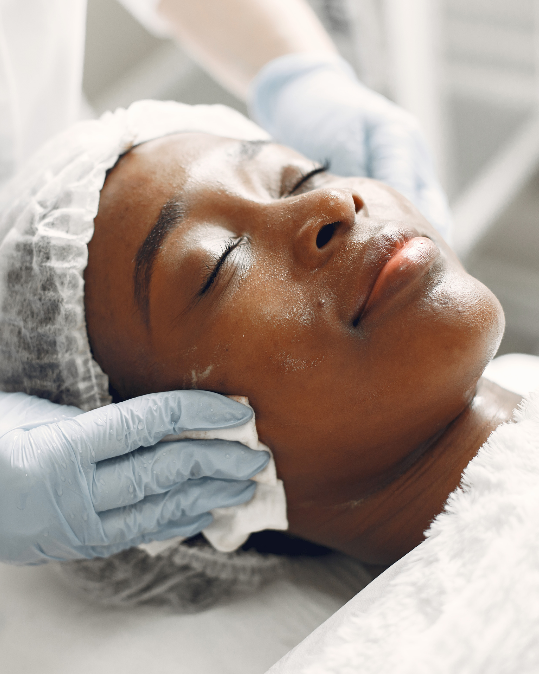 Close-up of a woman receiving a facial treatment, lying with her eyes closed, wearing a hair cap, with a skincare professional in gloves gently cleaning her face.