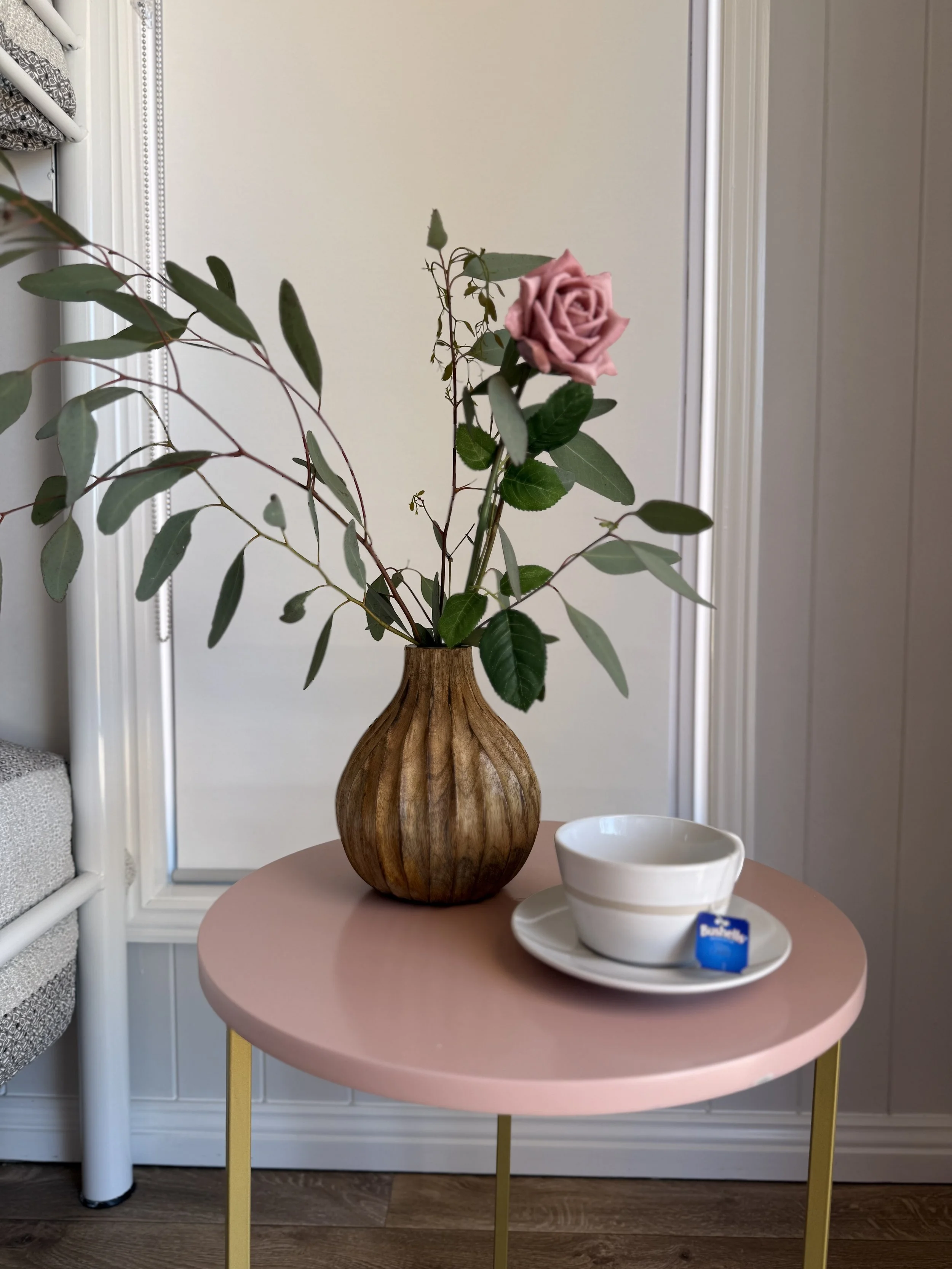 Pink tabletop with a brown wooden vase holding a pink rose and green foliage, and a white teacup and saucer with a tea bag label.