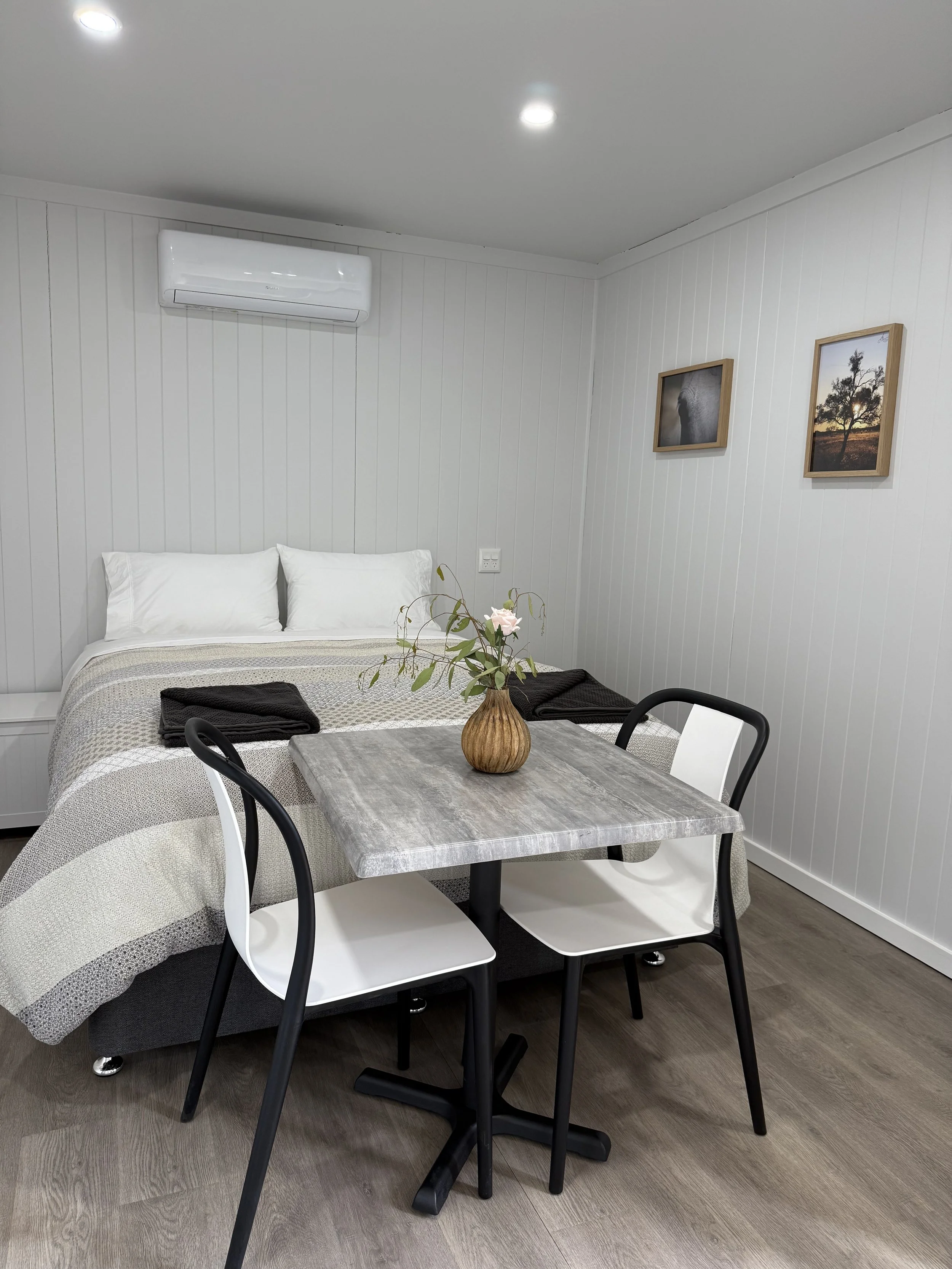 A bedroom with white paneled walls, a bed with white pillows, a gray and beige blanket, and black and white towels. There is a small dining table with a vase of flowers and four black and white chairs. A wall-mounted air conditioner and framed pictures are visible.