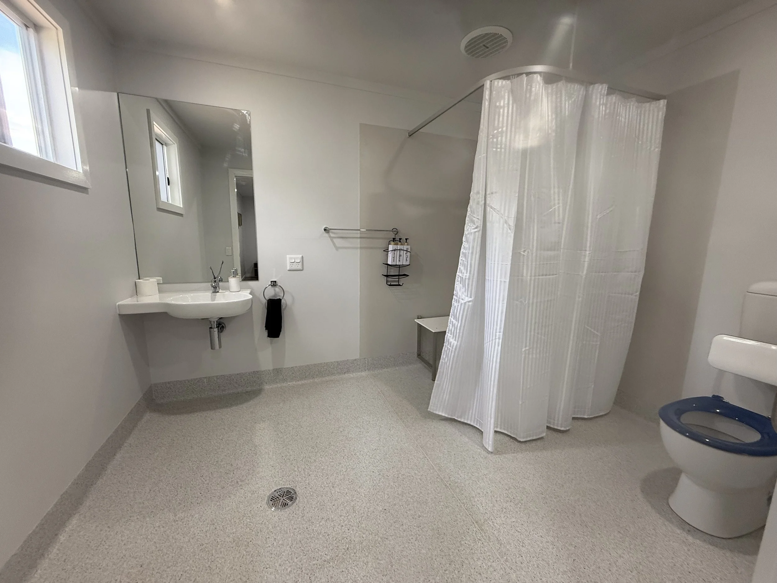 A clean, spacious bathroom with a white sink, large mirror, small windows, a toilet with a blue seat, a white privacy shower curtain, and a small stool.