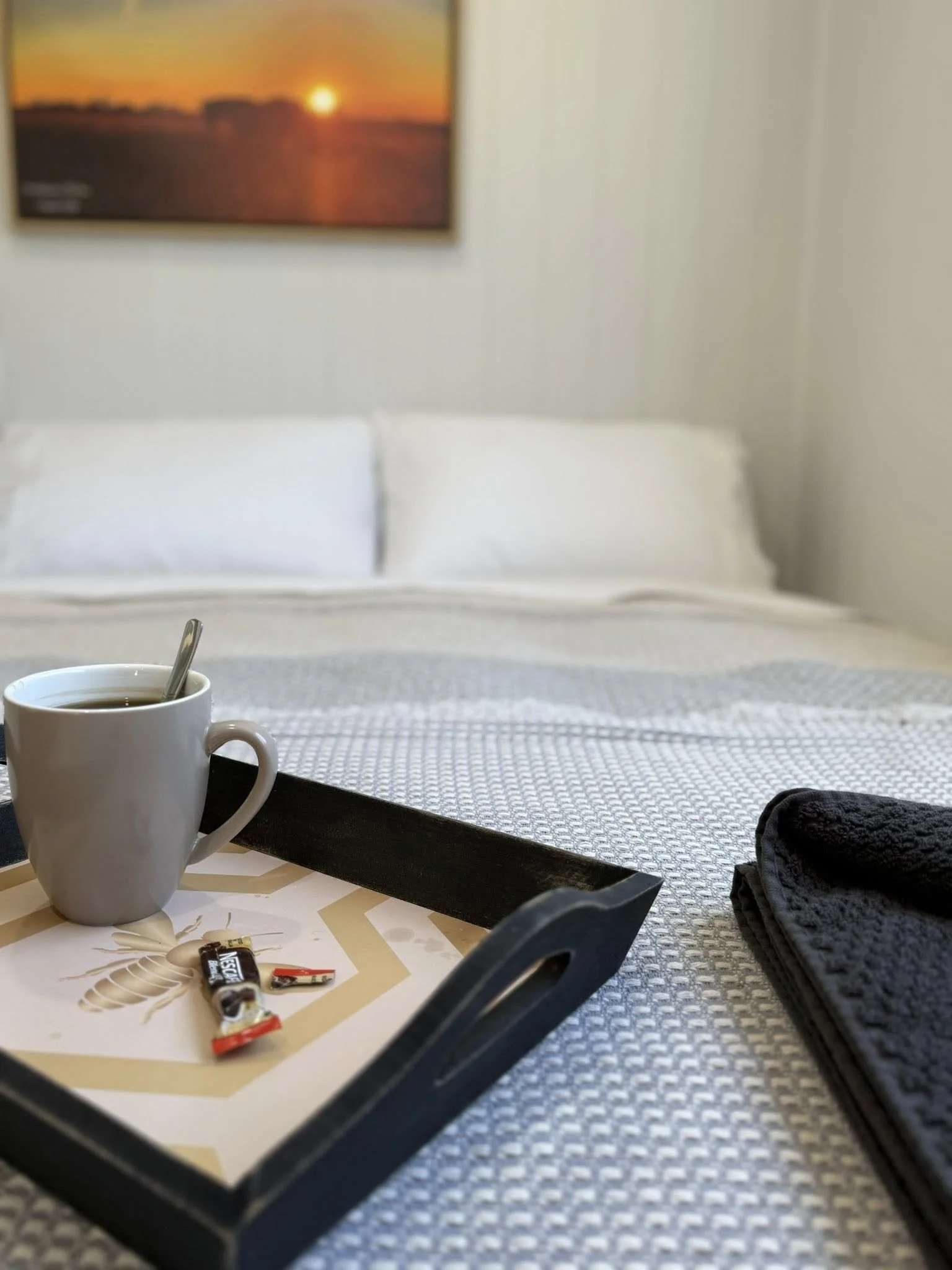 A hotel room with a bed, a tray with a coffee mug, and snacks, with a sunset picture on the wall.