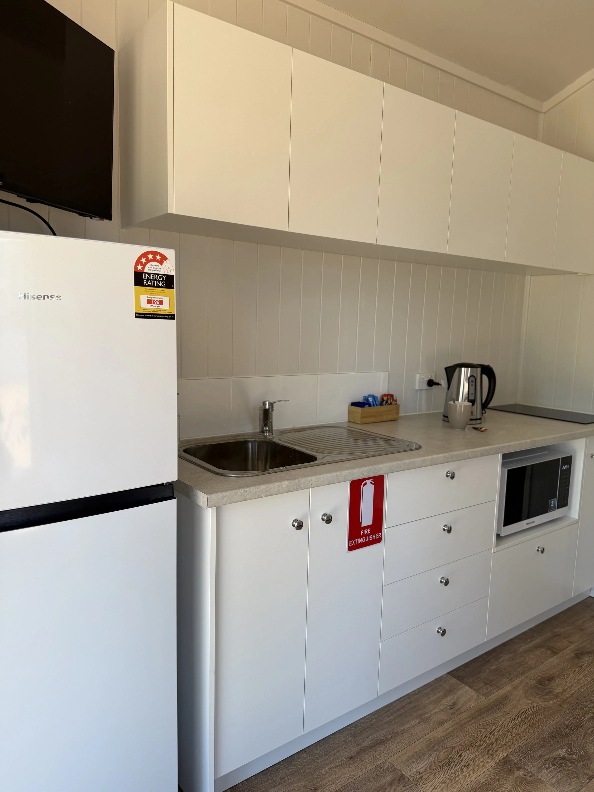 A compact kitchen with white cabinets, a mini refrigerator, a wall-mounted TV, a small sink, a microwave, a kettle, and a fire extinguisher sign on the cabinet door.