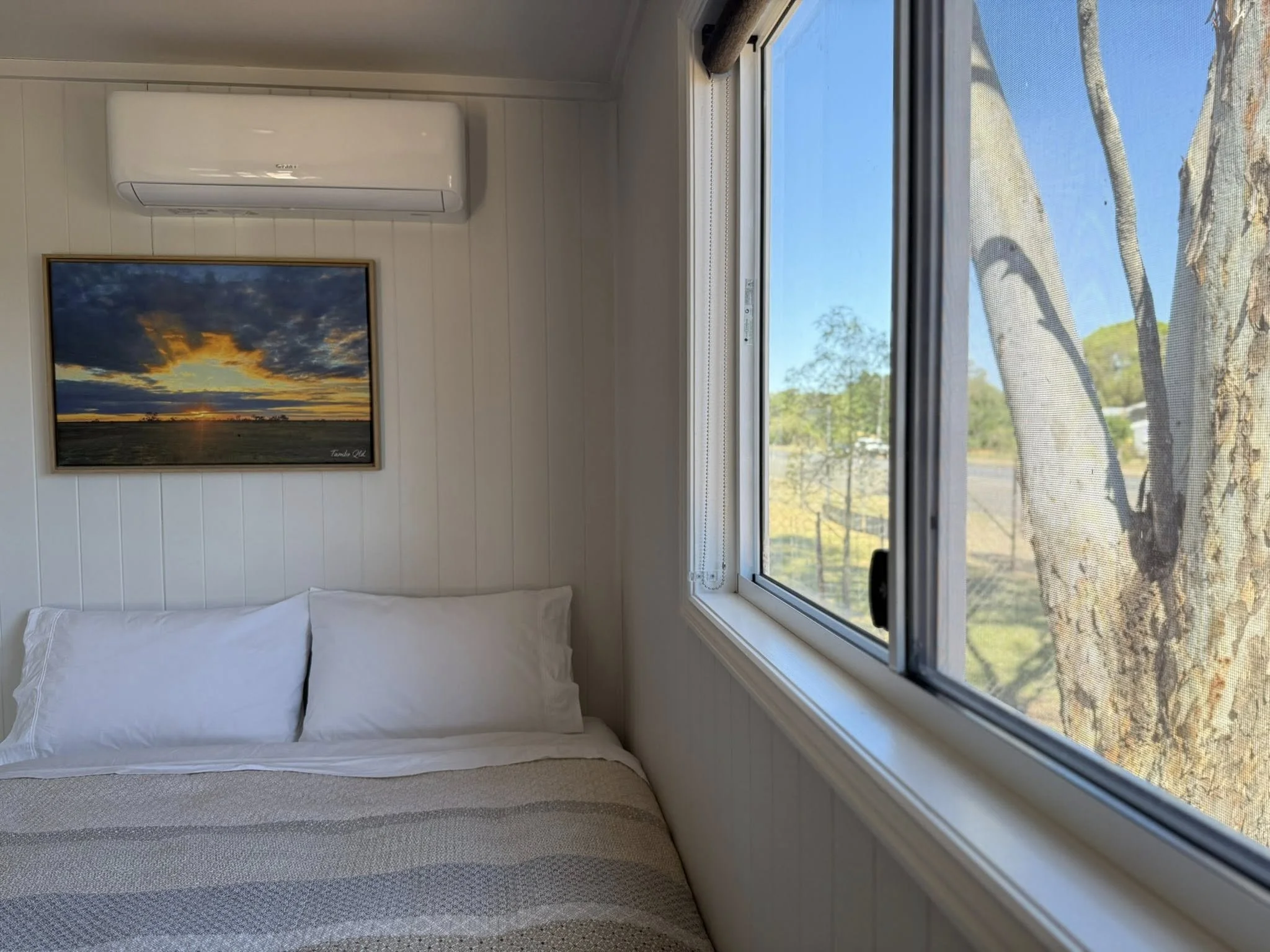 A cozy bedroom with white wood-paneled walls, a bed with white pillows and a beige striped blanket, a window showing trees and blue sky outside, a ceiling-mounted air conditioner, and a framed landscape photograph of a sunset over a field.