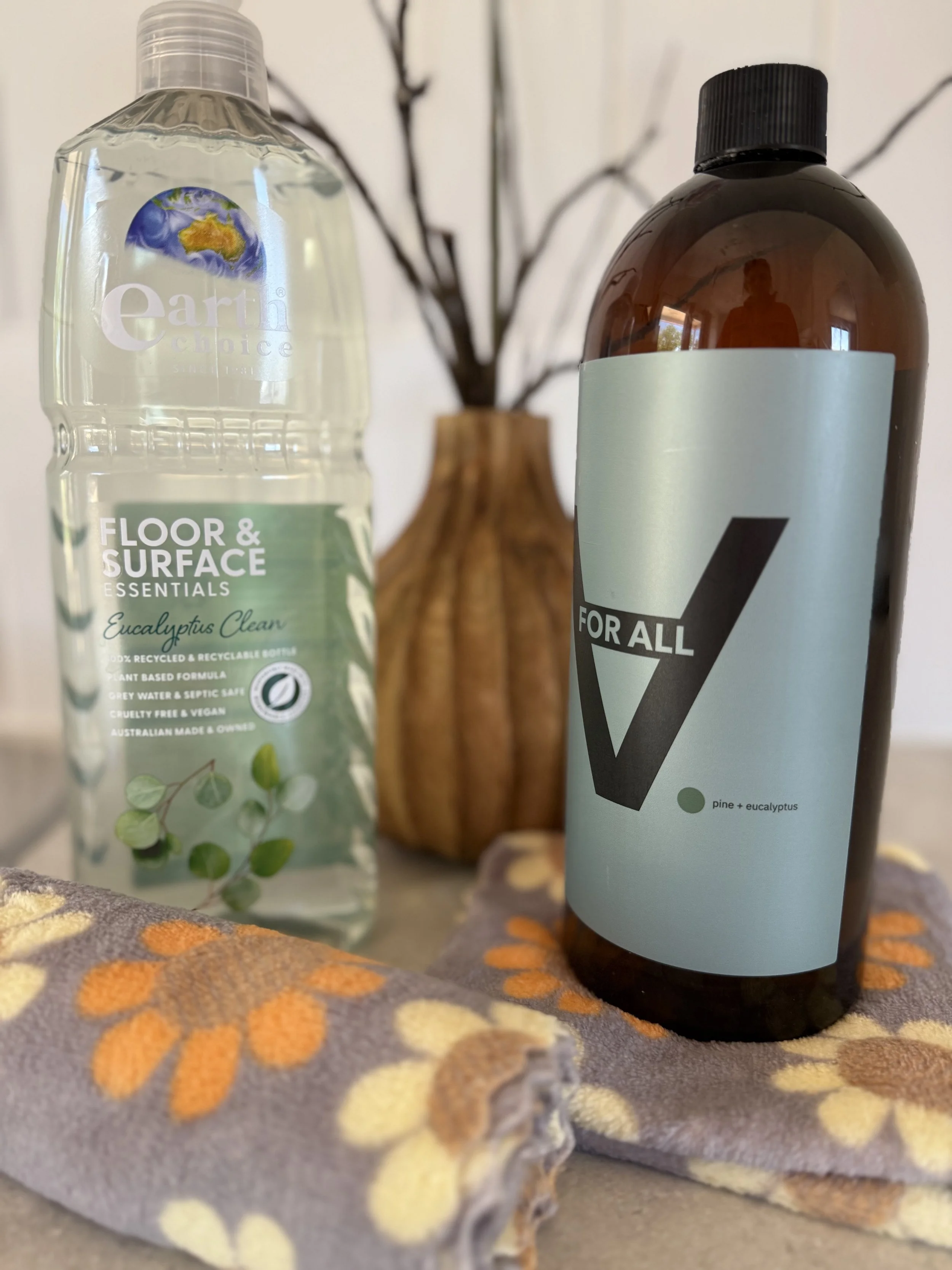 A bottle of Earth Choice Floor & Surface Essentials eucalyptus cleaner, a brown bottle with a label reading 'For All,' a decorative vase with dried branches, and a folded cloth with a floral pattern, all placed on a surface.
